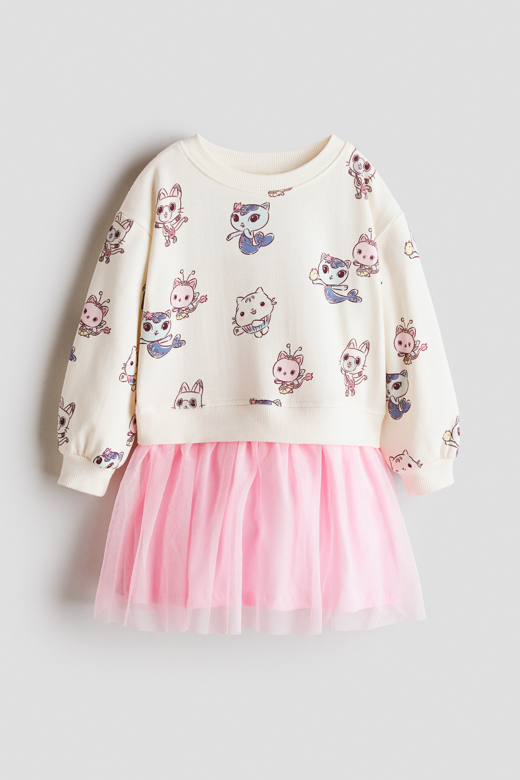 View larger image: A cream-coloured dress shown from the front, featuring a sweatshirt-style top with an all-over cartoon cat print, including some mermaid cats and some with wings, a ribbed crew neck, long sleeves with ribbed cuffs, and a ribbed hem, paired with an attached multi-layered pink tulle skirt.