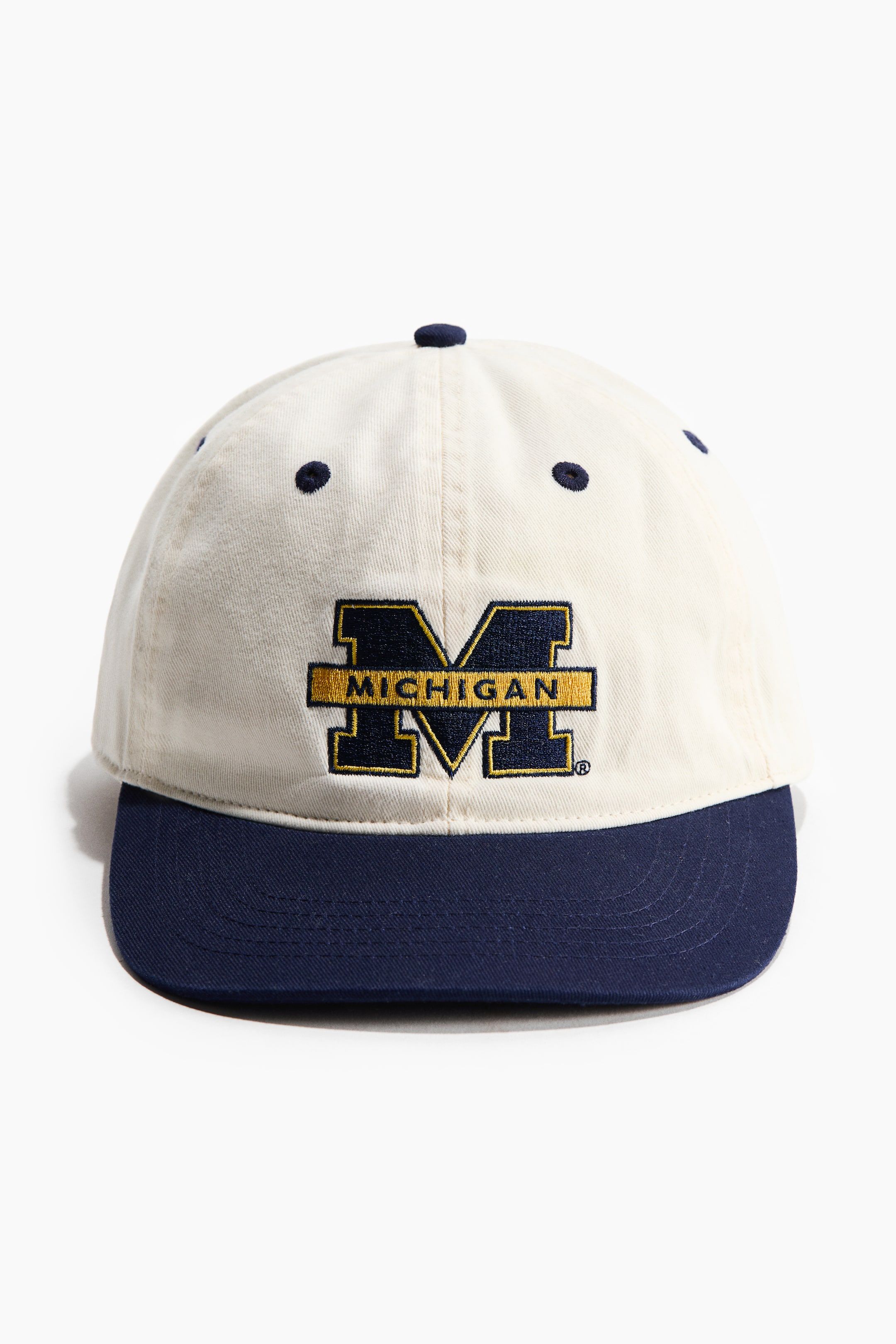 View larger image: The baseball cap is displayed front-facing, showcasing a cream crown accented by navy blue eyelets and a matching top button, complemented by a navy blue brim. Prominently featured on the front of the crown is a large, embroidered navy blue "M" outlined in gold, accompanied by the "MICHIGAN" text.