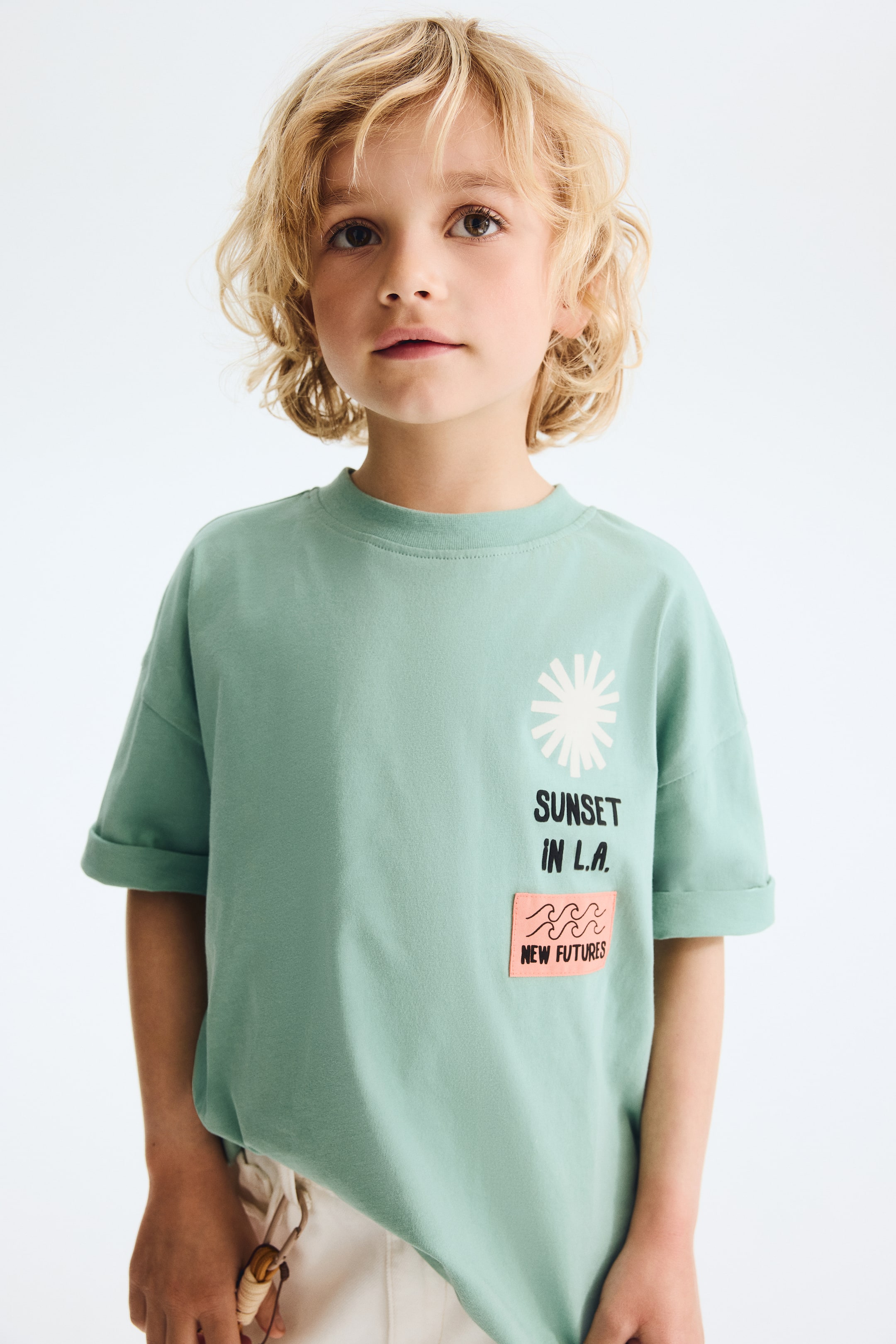 Visualizza l'immagine ingrandita: Una t-shirt verde chiaro è presentata su un bambino, leggermente girato verso destra, mettendo in risalto la vestibilità rilassata e le maniche corte arrotolate. Presenta una grafica a raggiera bianca, testo nero "SUNSET IN L.A." e una toppa rettangolare rosa con linee ondulate nere e testo "NEW FUTURES" sul davanti.