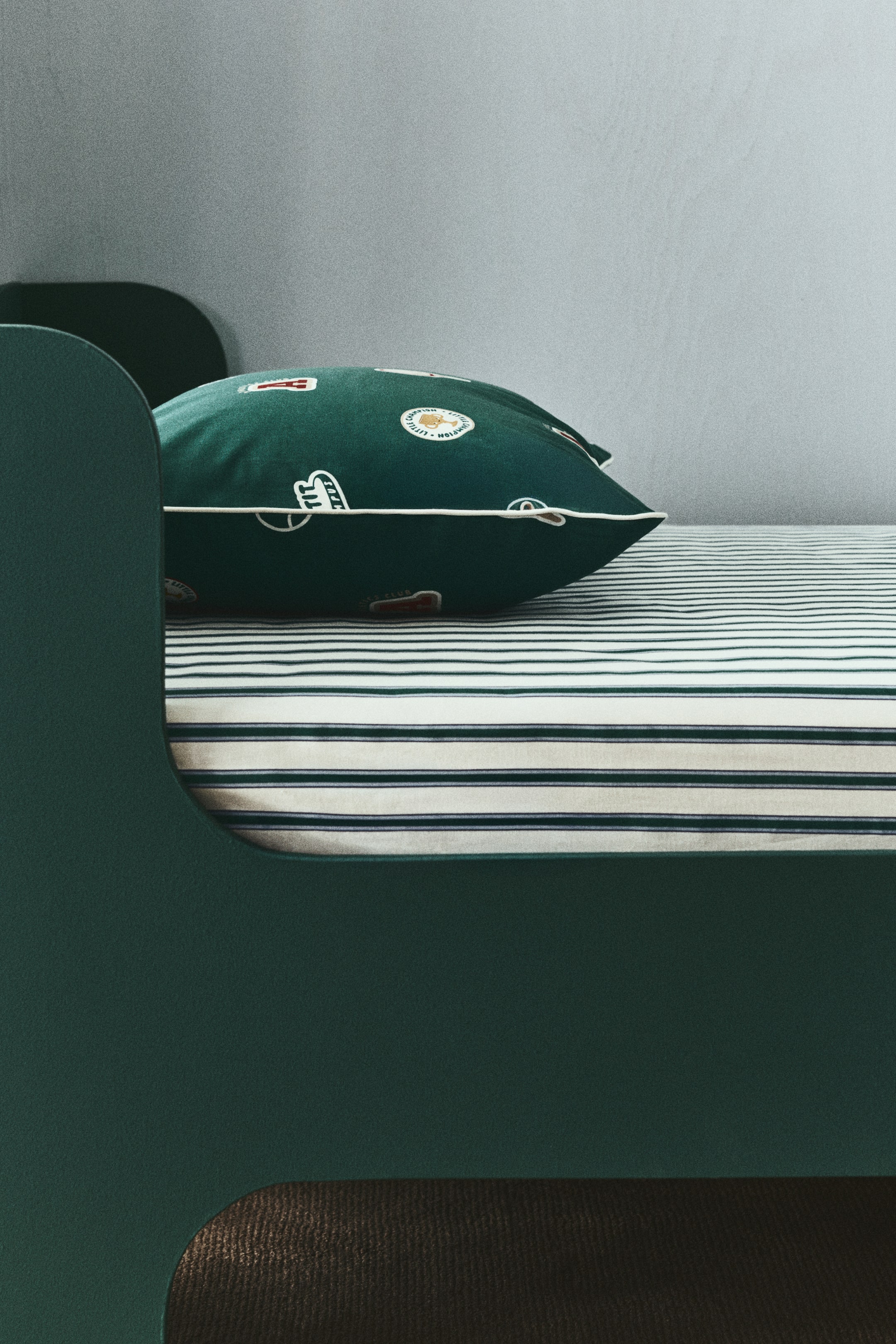 View larger image: A fitted sheet in a patterned cotton weave smoothly covers a mattress, extending towards the front. It features alternating white and dark green horizontal stripes of varying thickness, with a dark green pillow resting on its surface.