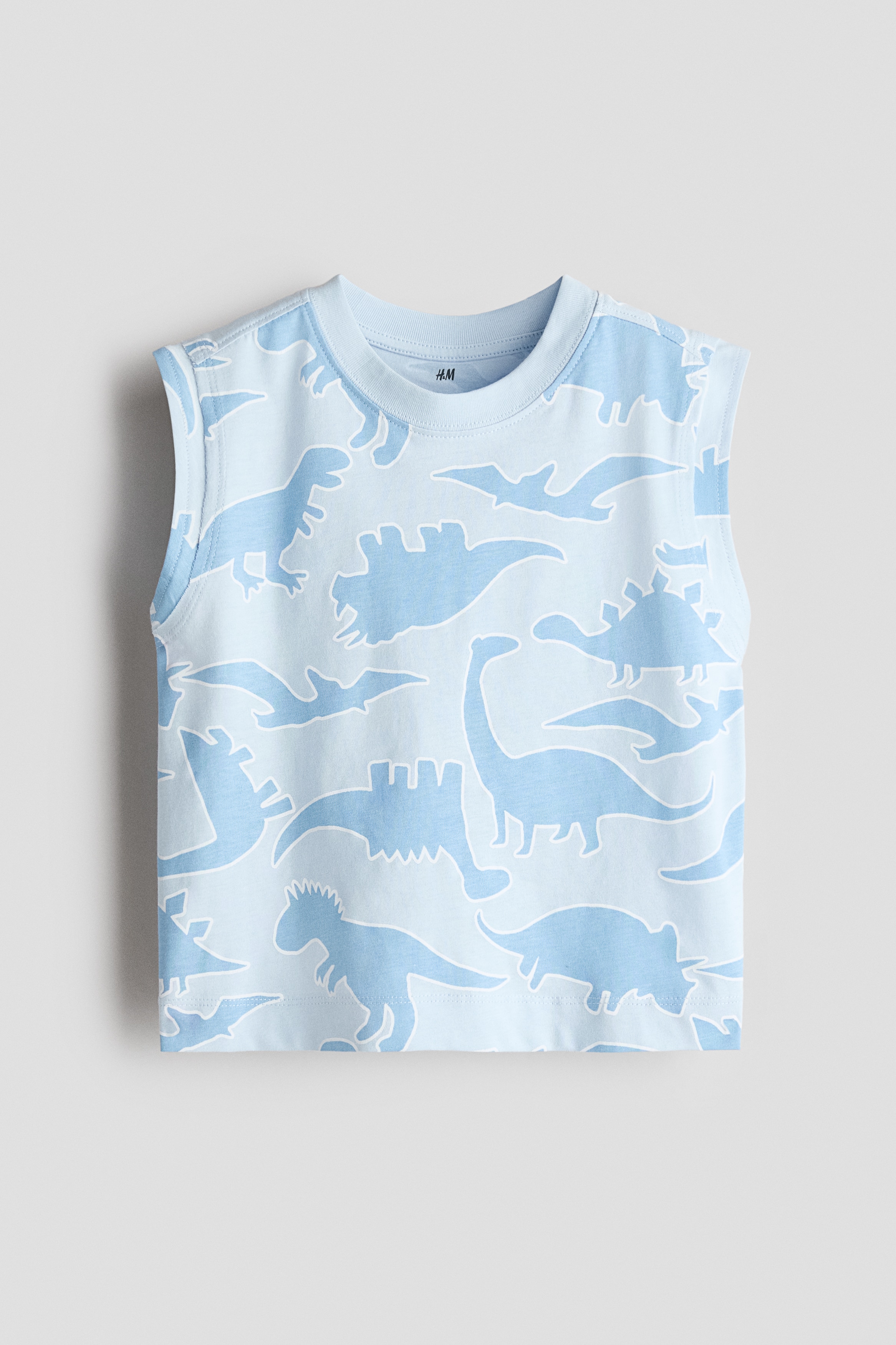 A light blue sleeveless tank top featuring an all-over dinosaur print and a round, rib-trimmed neckline is being worn by the model. The product is paired with matching dinosaur-print shorts and beige sandals.