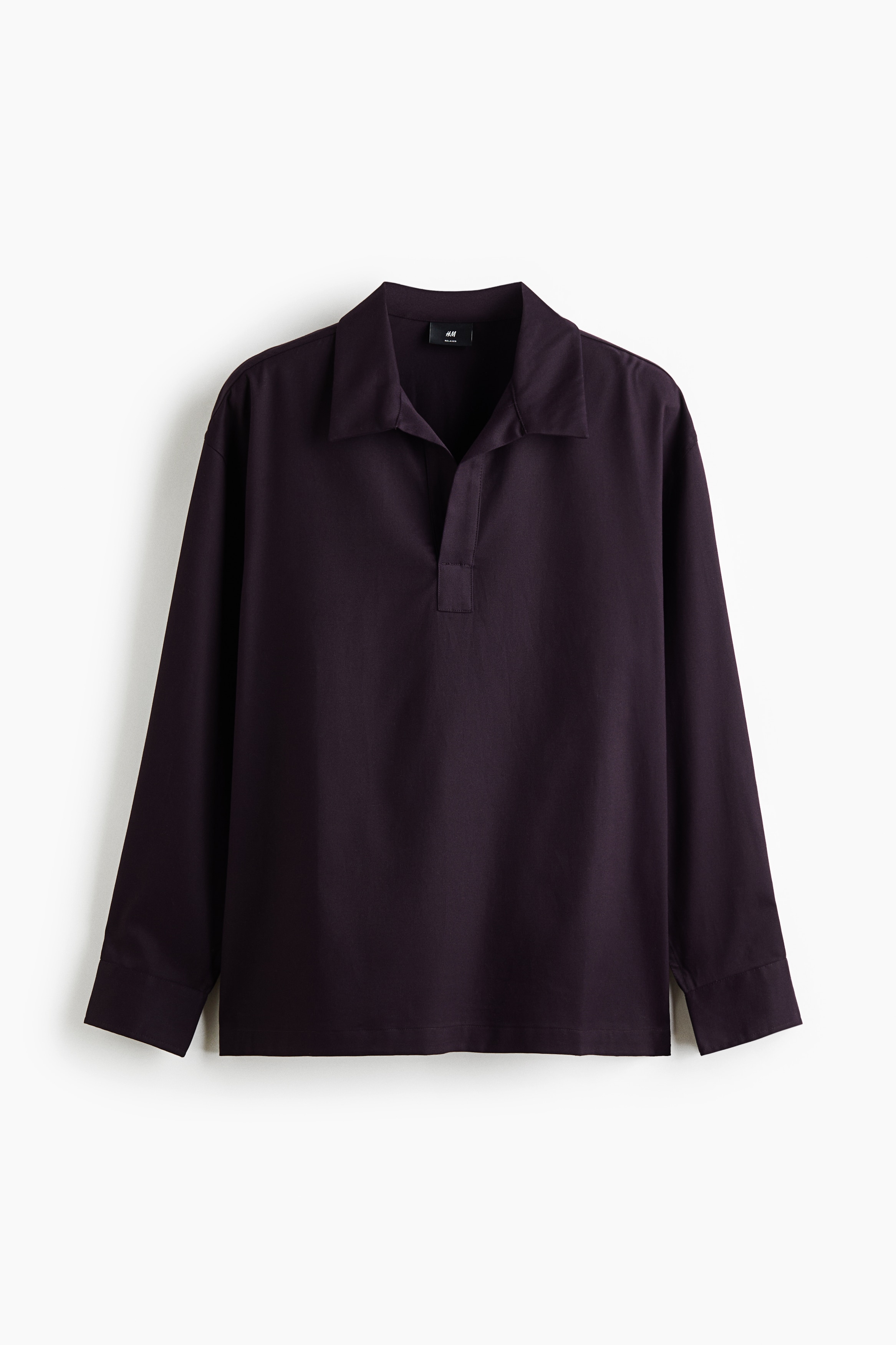 The model is wearing a dark purple twill popover shirt, featuring a relaxed fit, an open collar, and dropped shoulders. The shirt is styled with off-white trousers and a black tote bag.
