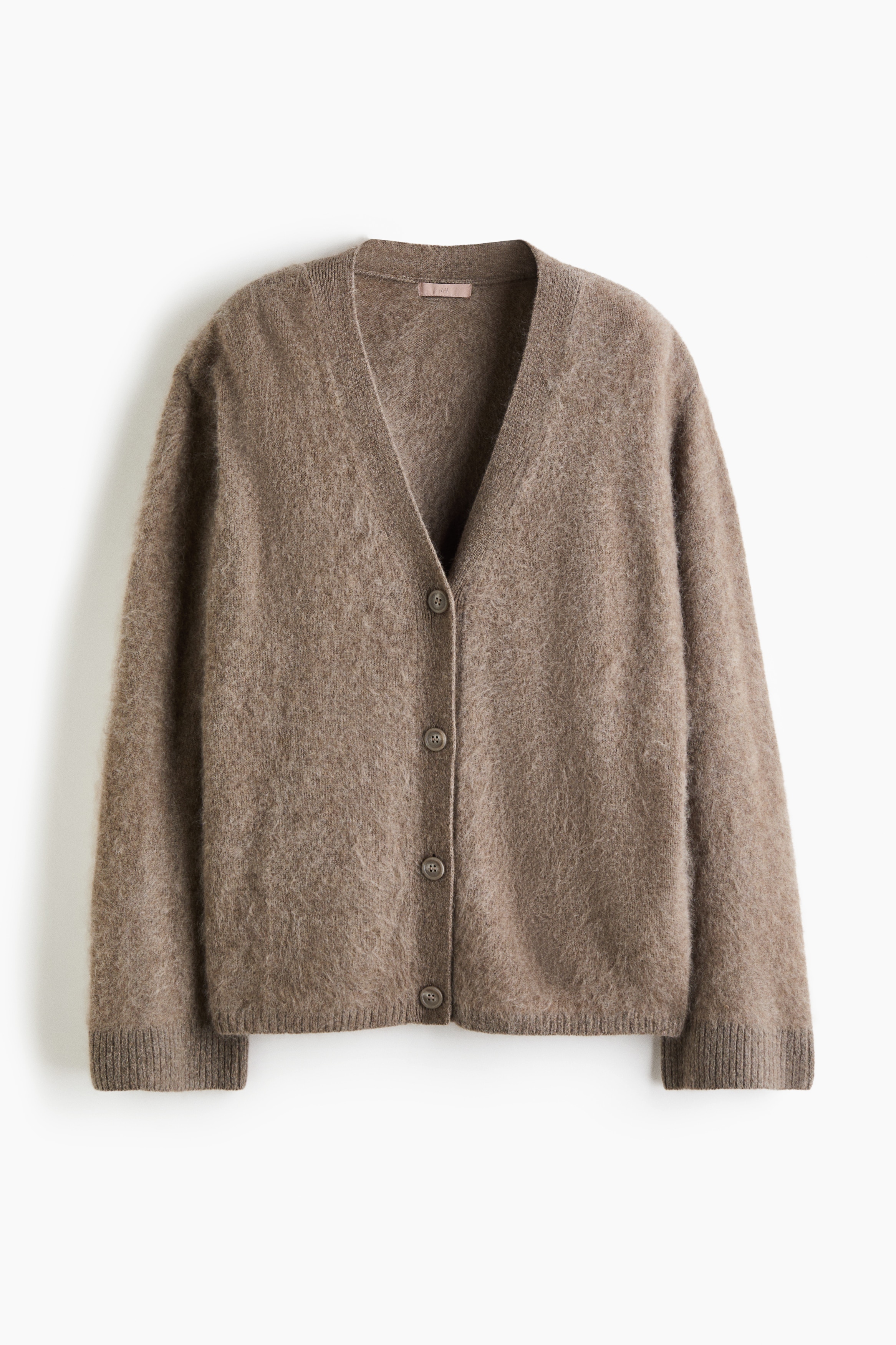An oversized, taupe-colored knit cardigan featuring a V-neckline, button closures, dropped shoulders, and ribbed cuffs is being worn by the model over a white bodysuit. The model is standing with crossed arms, holding one sleeve of the soft-textured cardigan.
