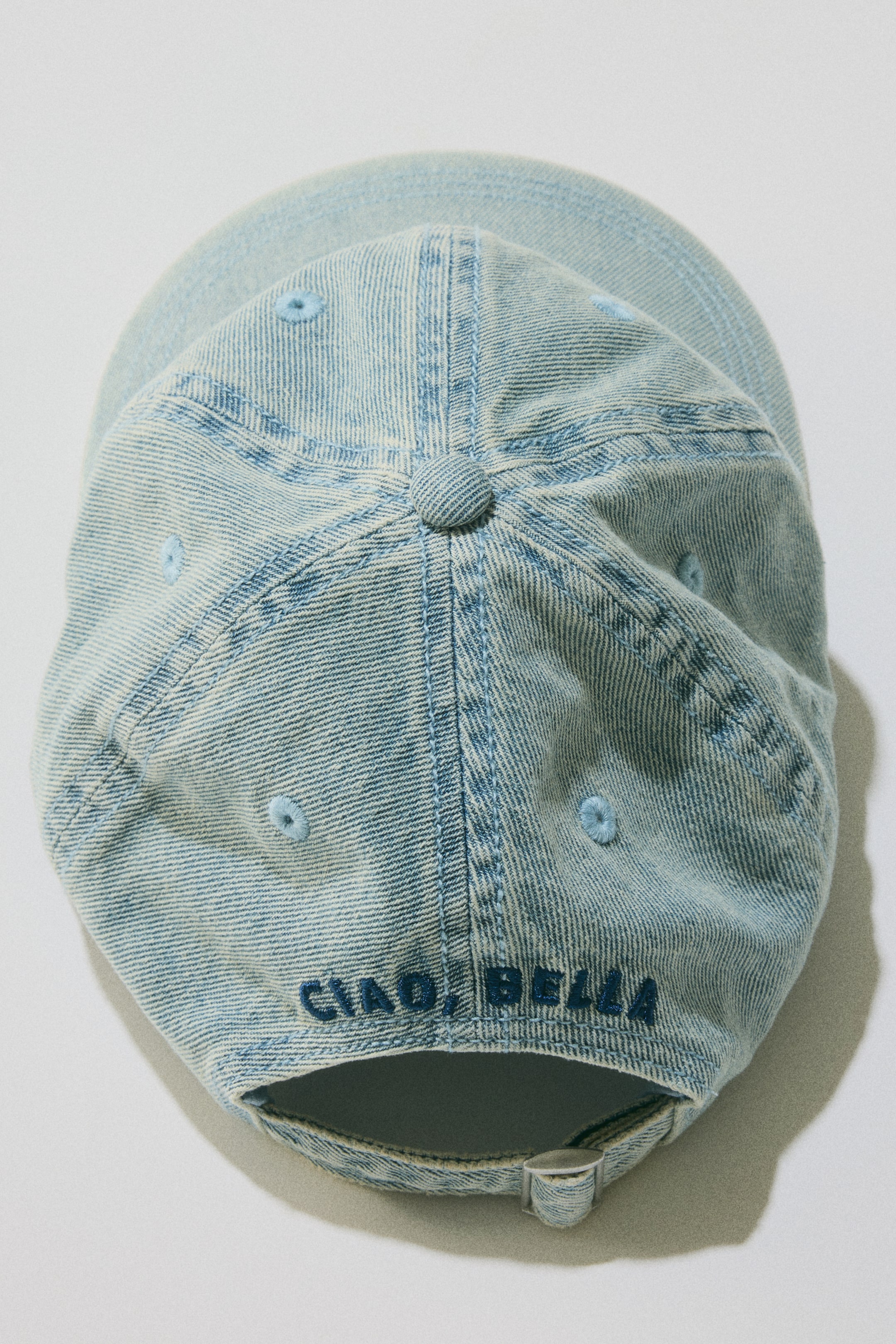 View larger image: A cap in light blue washed cotton twill is showcased from a top-down, rear angle. It features six panels, light blue embroidered eyelets, and "CIAO, BELLA" embroidered in dark blue across the back, with an adjustable tab and metal slide buckle visible at the rear.