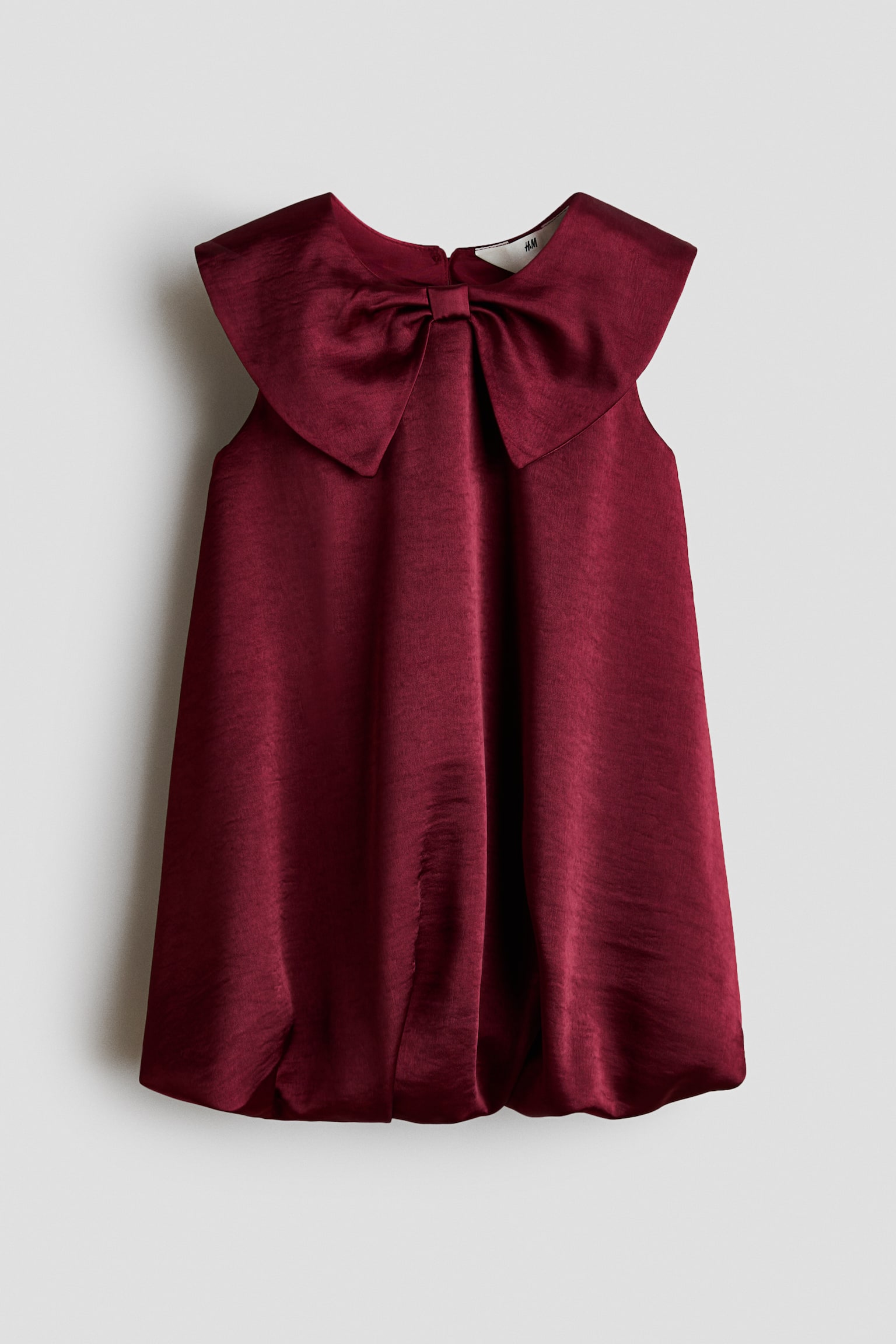 A sleeveless burgundy dress made of a shiny woven fabric is hanging front-facing, showcasing an oversized bow-shaped collar at the neckline. The dress drapes into a flared silhouette and finishes with a gathered bubble hem.