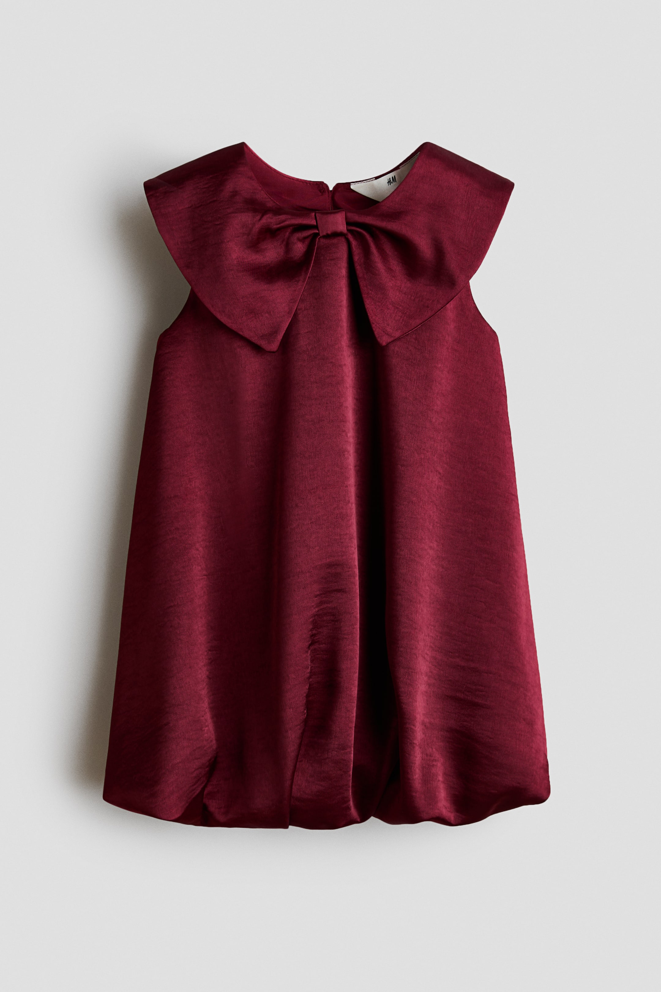 View larger image: A sleeveless burgundy dress made of a shiny woven fabric is hanging front-facing, showcasing an oversized bow-shaped collar at the neckline. The dress drapes into a flared silhouette and finishes with a gathered bubble hem.