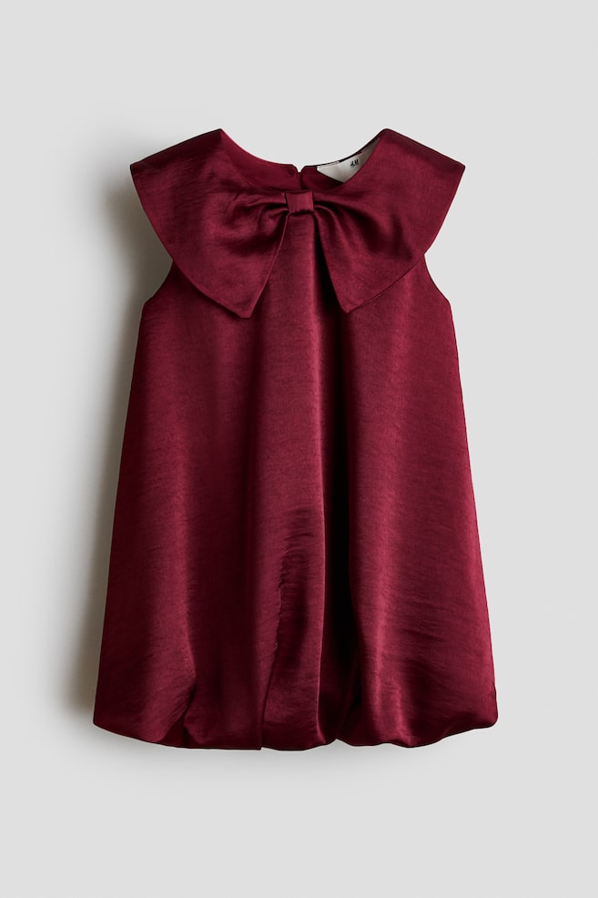 A sleeveless burgundy dress made of a shiny woven fabric is hanging front-facing, showcasing an oversized bow-shaped collar at the neckline. The dress drapes into a flared silhouette and finishes with a gathered bubble hem. - 1