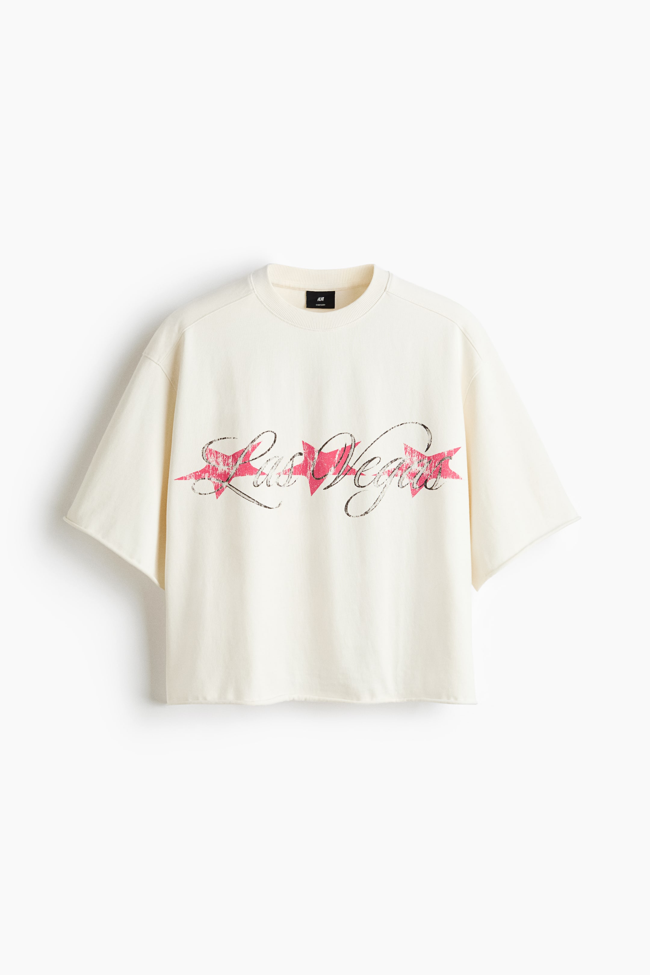 View larger image: A cream T-shirt is displayed front-facing, featuring a ribbed crew neck and a distressed "Las Vegas" graphic print in black over pink stars and a banner across the chest. It is designed with wide, dropped-shoulder short sleeves and a cropped hem, both with raw edges.