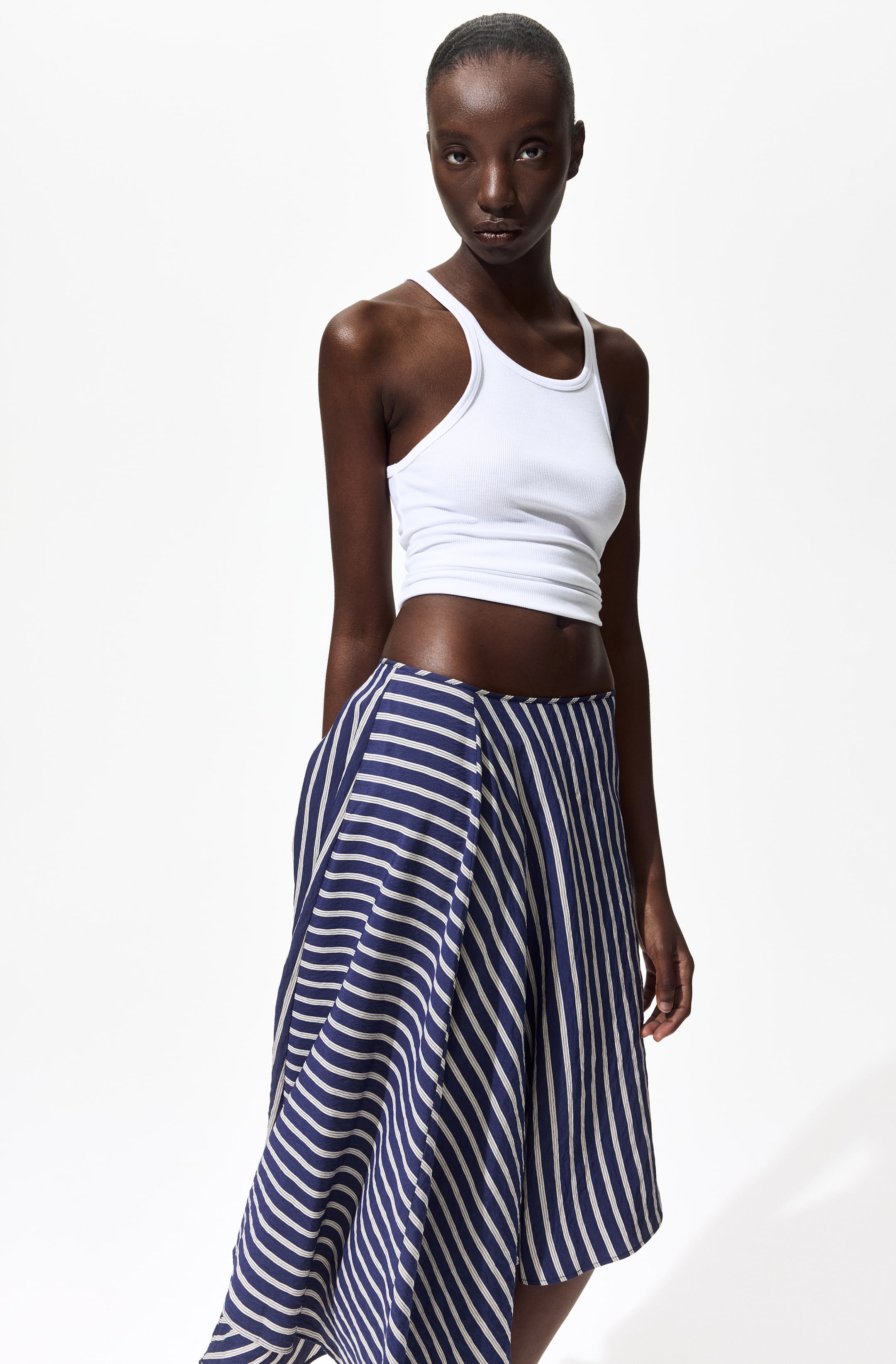 Visualizza immagine più grande: La modella indossa una gonna a righe blu e bianche, lunga fino al ginocchio, caratterizzata da un bordo sottile in vita, una silhouette svasata e un orlo asimmetrico.