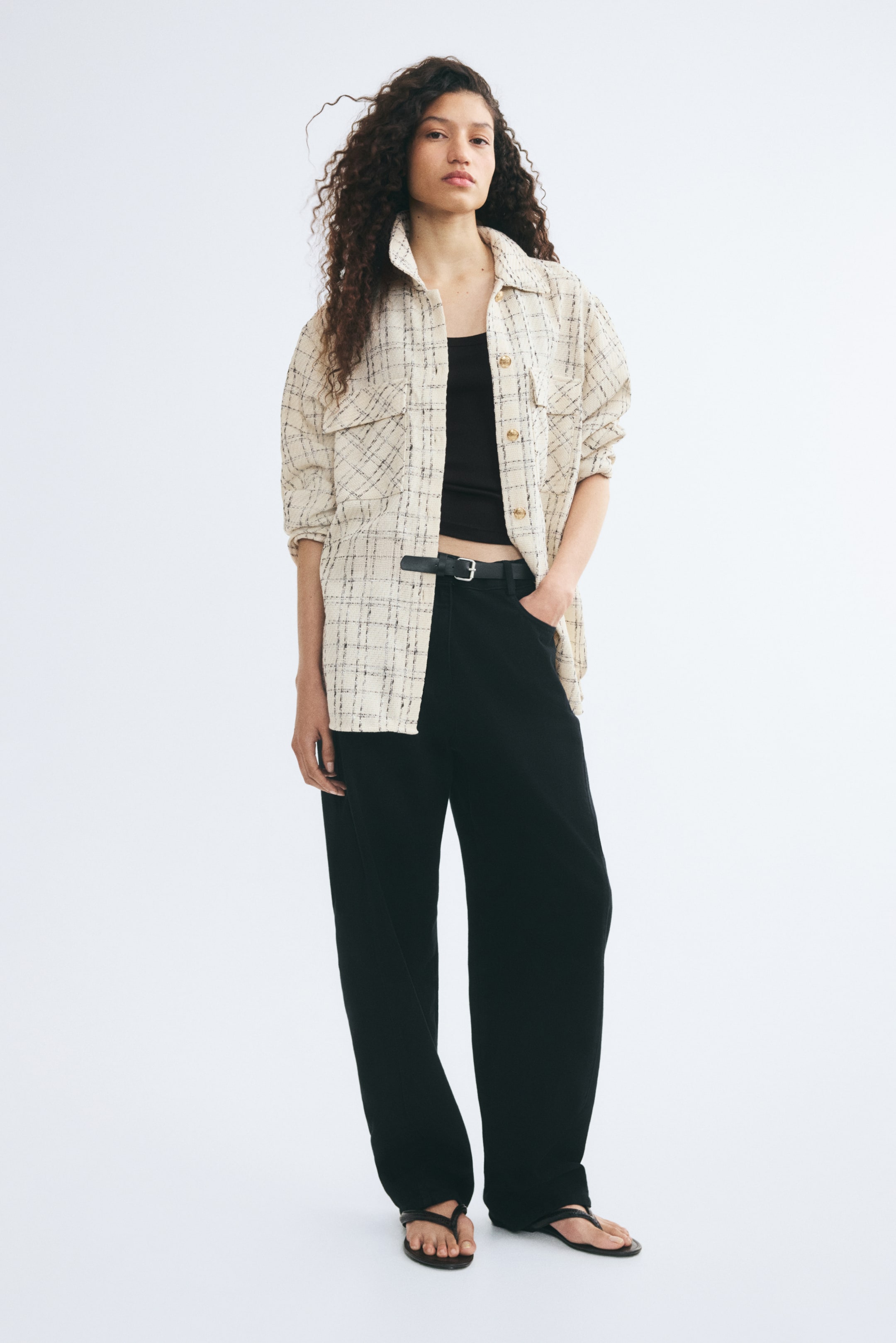 Visualizza l'immagine ingrandita: Uno shacket in tweed color crema con texture, caratterizzato da un motivo a griglia nero, tasche con patta sul petto e bottoni color oro, indossato aperto dalla modella. La camicia oversize è sovrapposta a un crop top nero e a pantaloni neri a gamba larga, stretti in vita con una cintura.