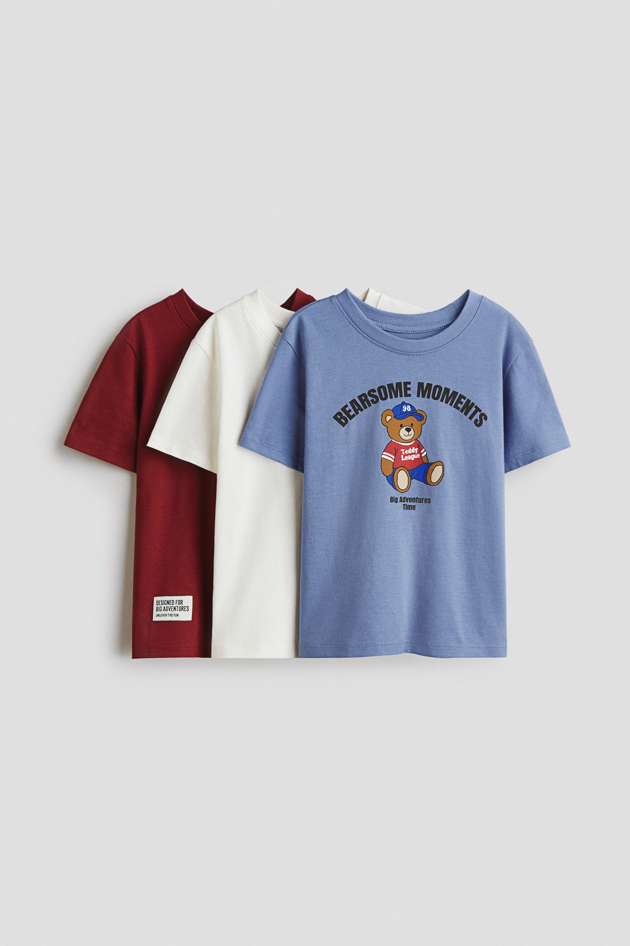 View larger image: Three classic T-shirts are shown from the front, each made from soft cotton jersey with a round, rib-trimmed neckline and a straight-cut hem. The blue T-shirt features a graphic print of a teddy bear wearing a cap and striped shirt, with “Bearsome Moments” text. The off-white and dark red T-shirts present plain designs, with the dark red one displaying a white fabric label on its lower left side.
