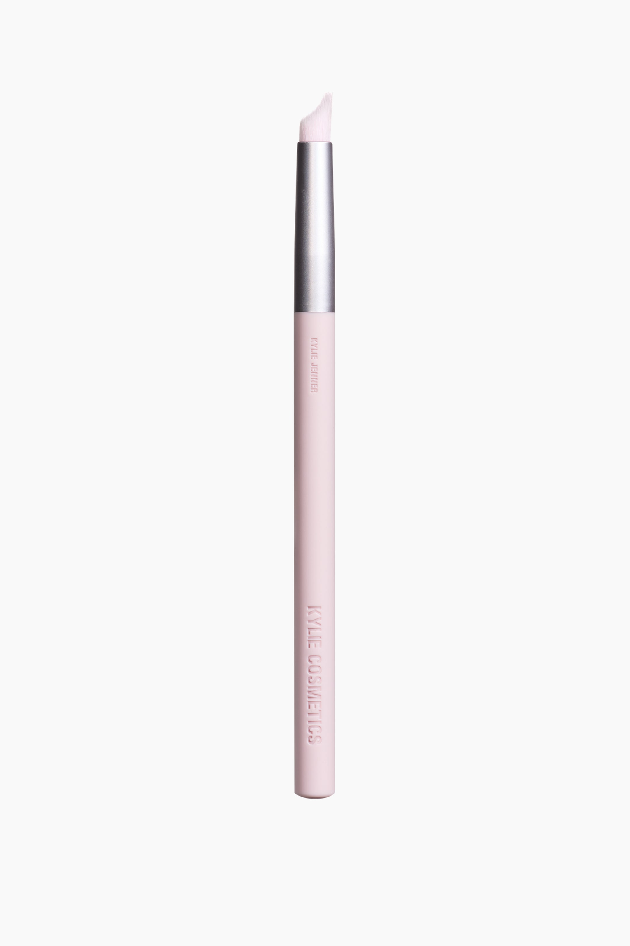 Se større billede: En lyserød makeupbørste står oprejst, med en sølvfarvet ferrule og hvide, vinklet syntetiske børstehår øverst. Skaftet viser "KYLIE JENNER" og "KYLIE COSMETICS" branding i præget skrift.