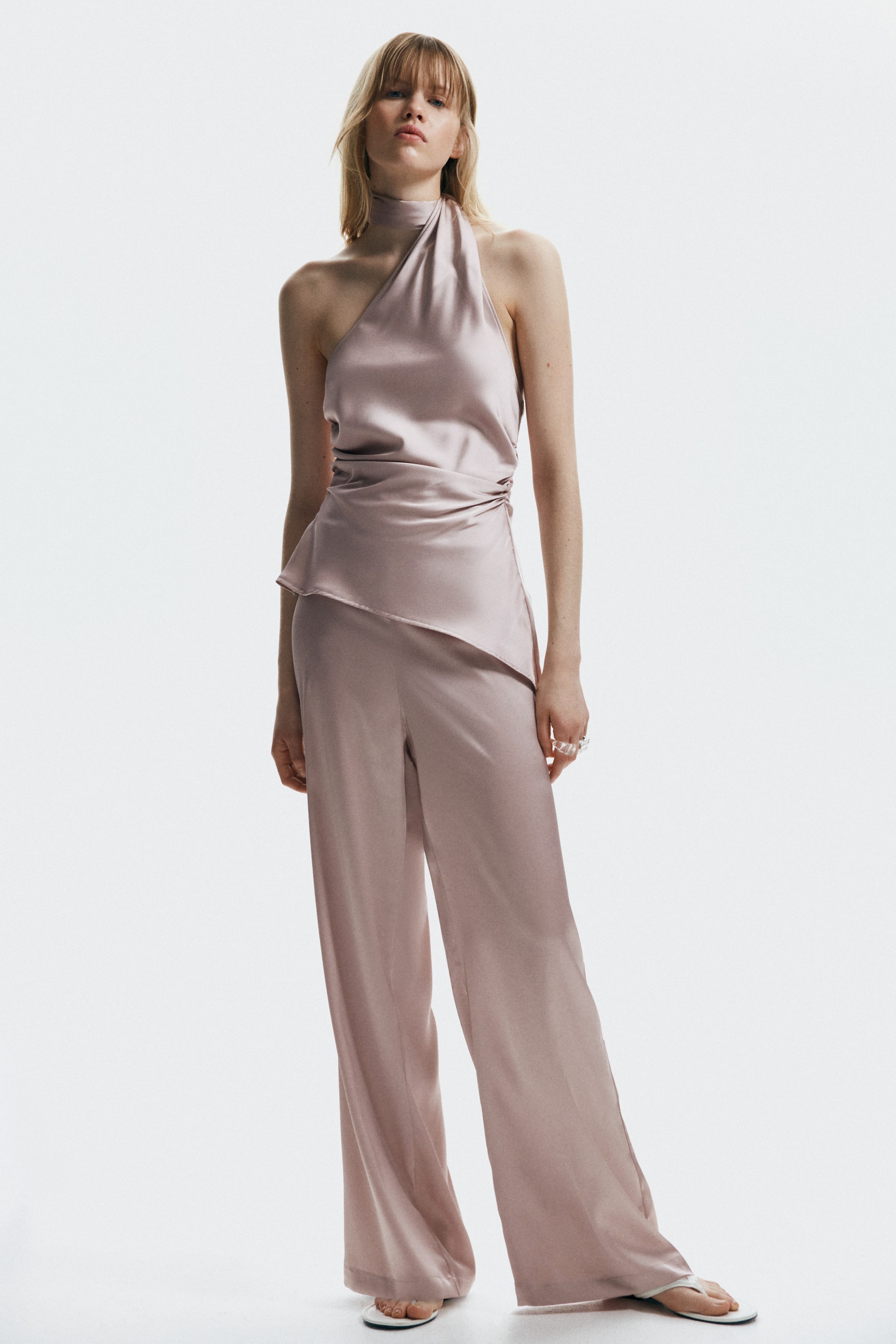 View larger image: A pale rose satin two-piece set is displayed, featuring a draped halter-neck top and wide-leg trousers. The top is cinched at one side of the waist, forming an asymmetric peplum-style hem that elegantly flows over the coordinating wide-leg trousers.