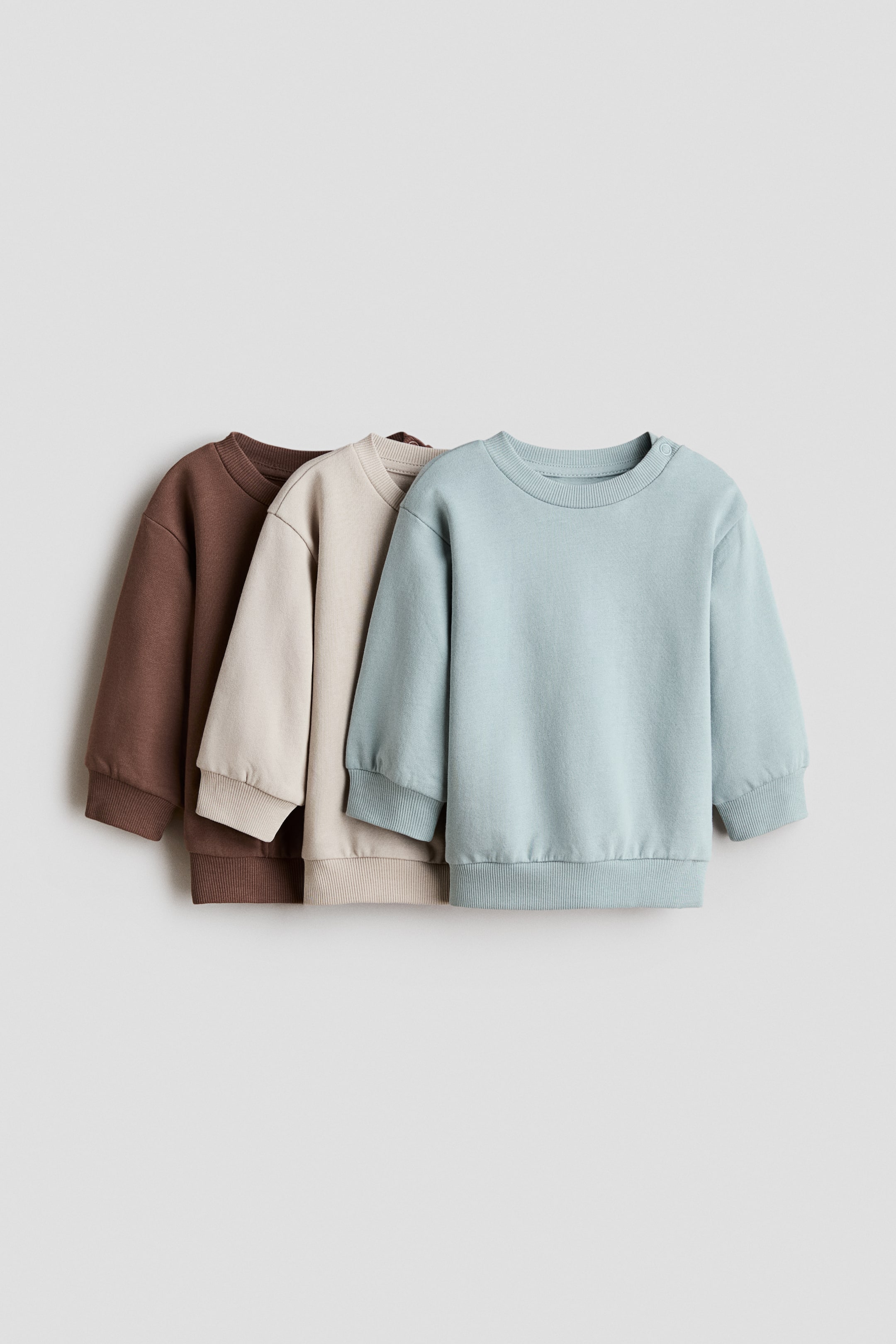 Se større billede: Tre baby-sweatshirts præsenteres i en lagdelt opstilling, let vinklet mod højre. En lyseblå sweatshirt ses forrest, med en beige bagved og en brun bagerst. Hver har rund hals, lange ærmer med ribkanter ved manchetterne og en ribkant forneden. Den lyseblå sweatshirt har desuden trykknapper på venstre skulder.