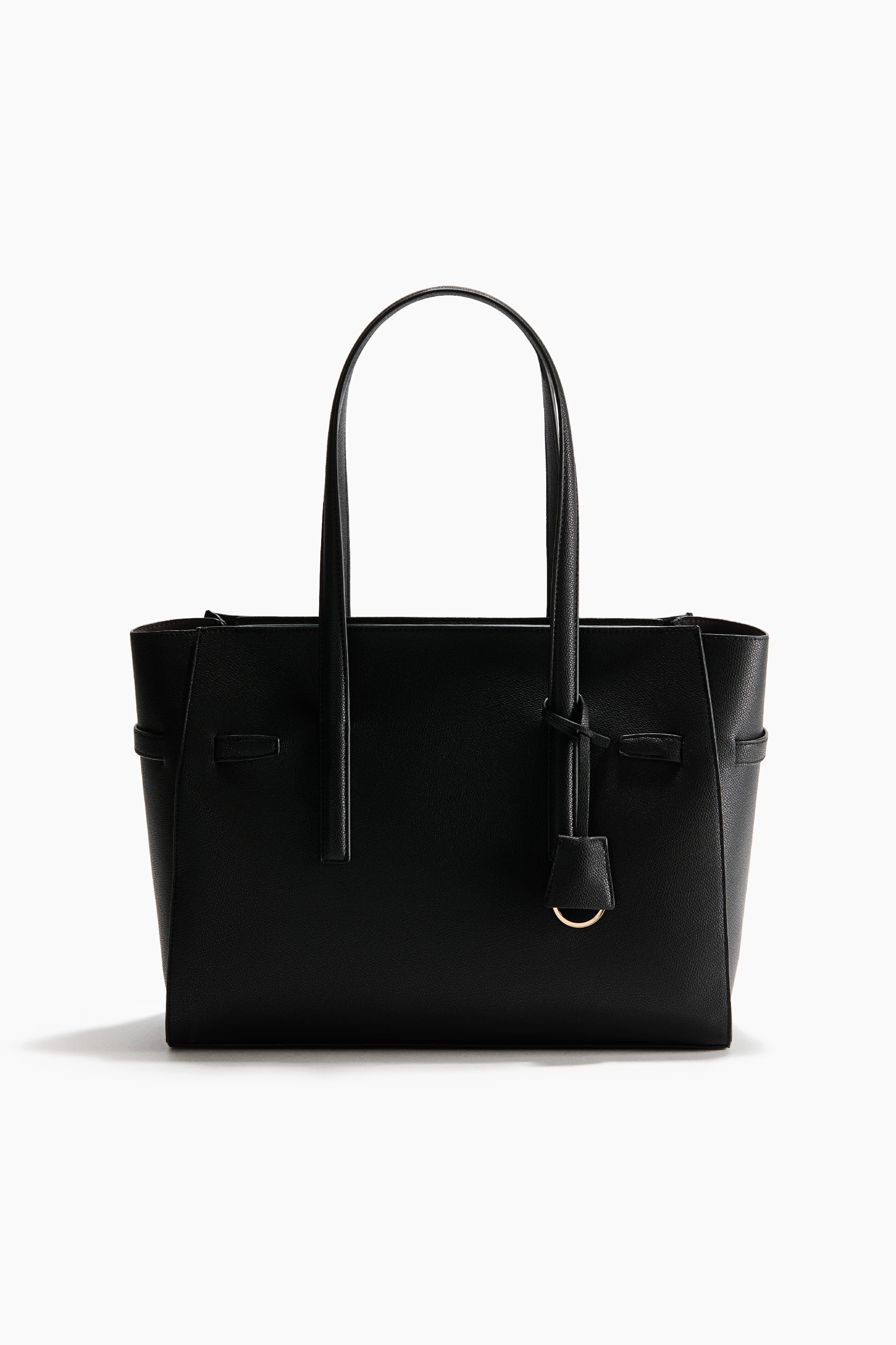 A structured black tote bag in textured coated fabric is being carried by the model, featuring long top handles and a decorative bag tag with a gold-tone ring. The bag is styled with a matching dark leather-look outfit and black heeled boots.