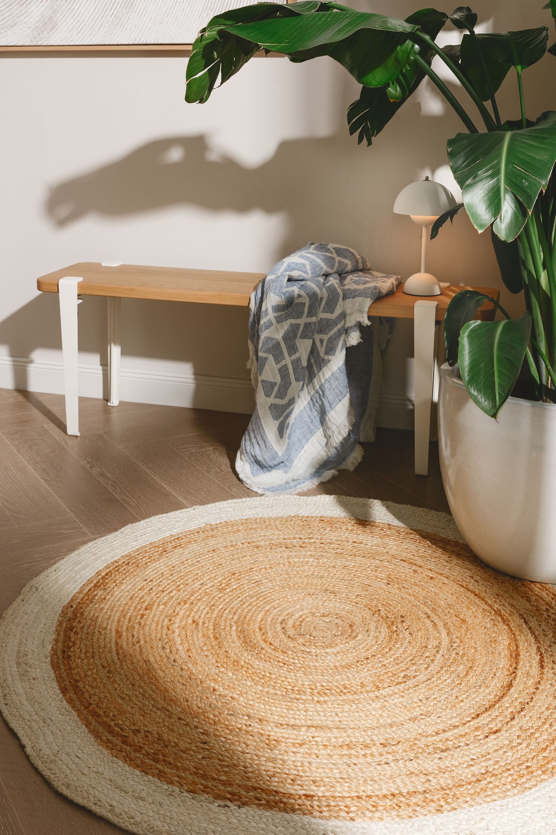 Se større billede: Et rundt tæppe fremviser et flettet jutedesign, med et naturligt lysebrunt centralt område, der danner koncentriske cirkler, og en kontrasterende hvid ydre kant.