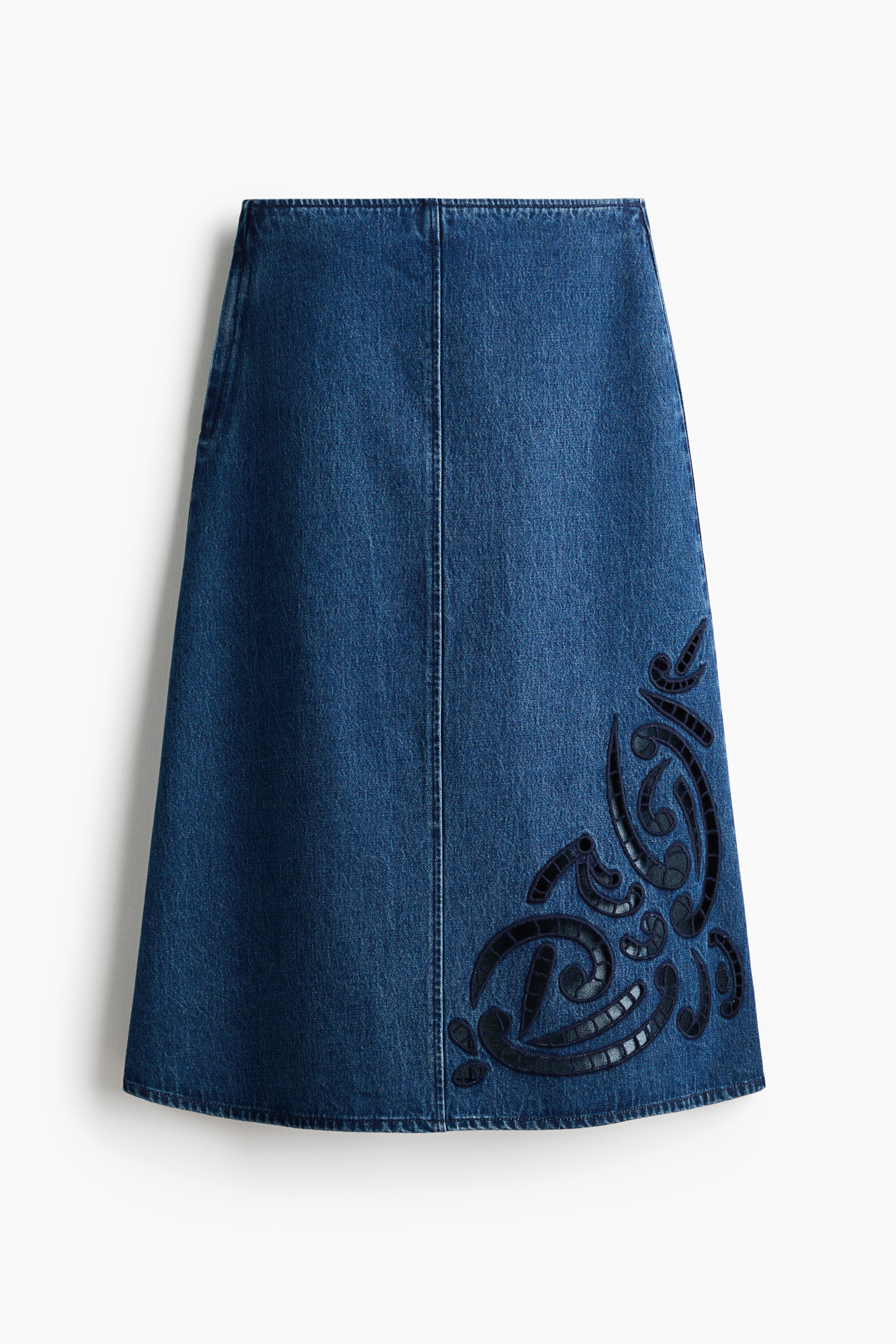 Ver imagen más grande: Una falda de línea A de mezclilla azul medio se presenta plana, de frente. Presenta una costura vertical visible en el centro frontal y un diseño intrincado de apliqué negro texturizado con patrones ondulantes en su lado inferior derecho.