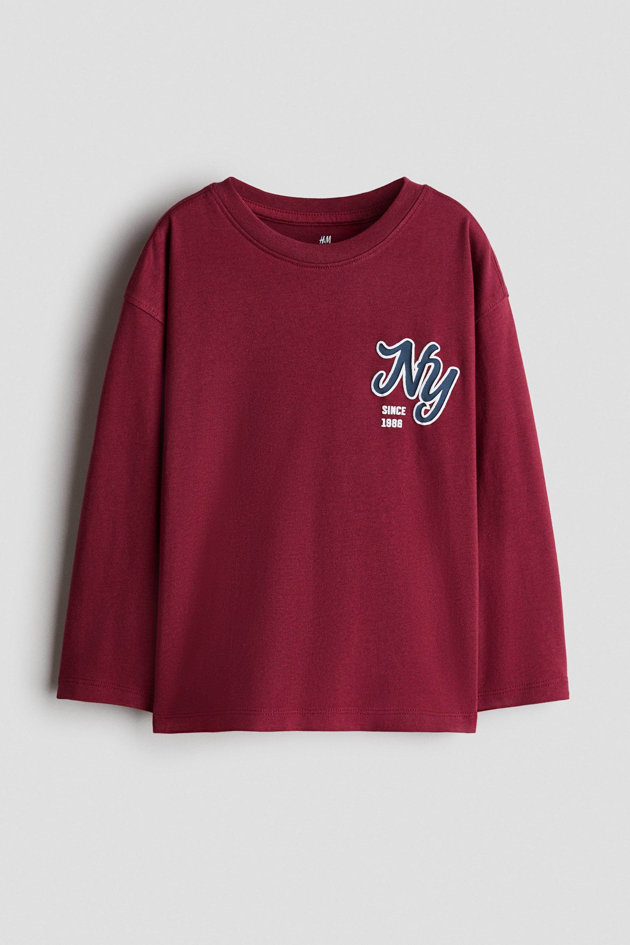 View larger image: A front-facing view shows a dark red, long-sleeve tee in cotton jersey featuring dropped shoulders and a ribbed round neckline. A small navy blue and white graphic of cursive "Ny" with the text "SINCE 1986" is printed on the left chest.