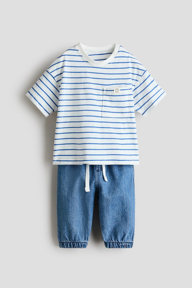 Ensemble bébé deux pièces présenté de face, composé d'un t-shirt blanc à rayures horizontales bleues, doté d'un col rond côtelé et d'une poche poitrine avec une petite étiquette. Le pantalon bleu effet denim présente une taille élastiquée avec cordons blancs et des bas de jambe élastiqués. - 1