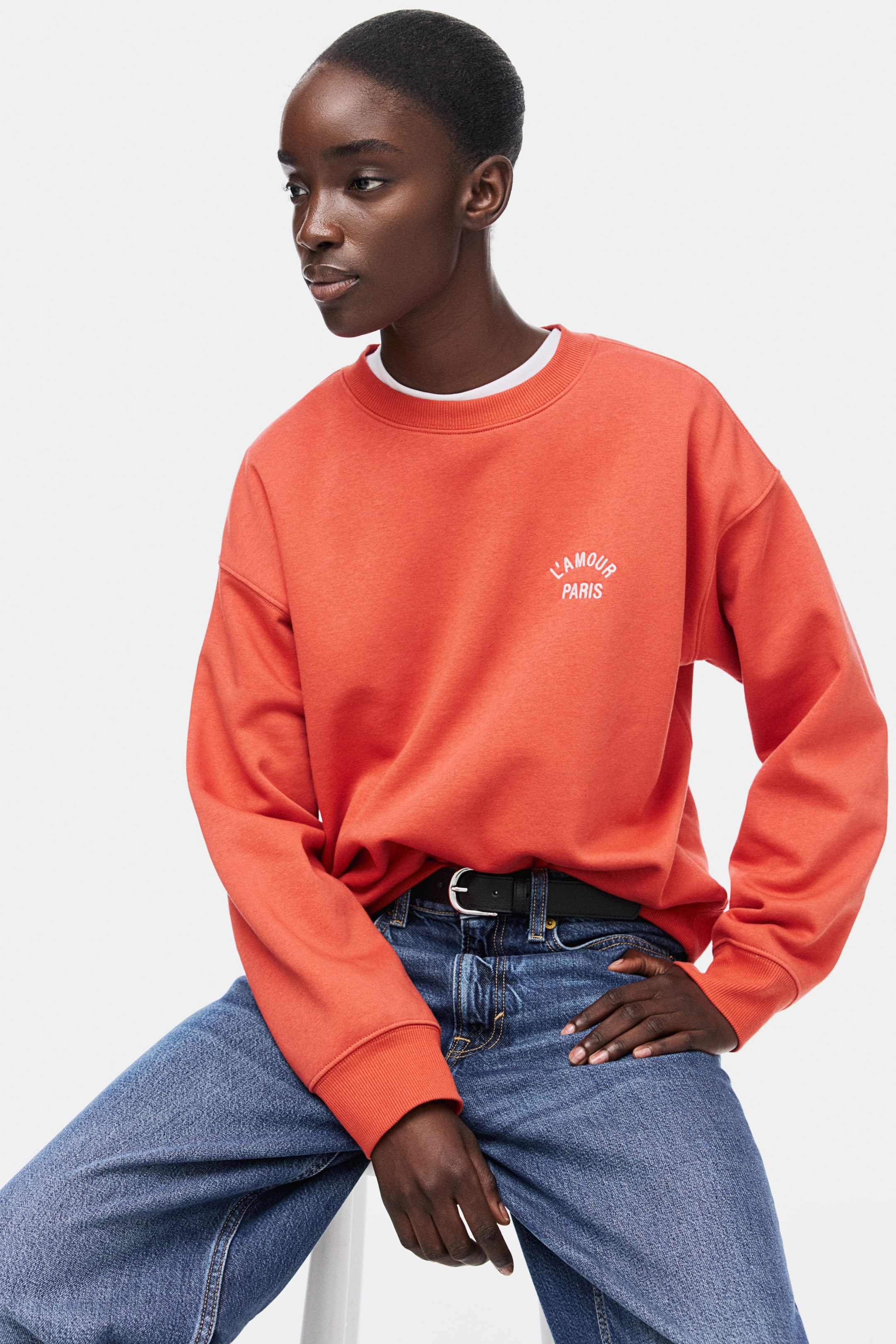 View larger image: The orange crewneck sweatshirt is showcased from a front-facing angle. It features a white "L'AMOUR PARIS" text design on the left chest, along with a ribbed crewneck, ribbed cuffs, and a ribbed hem, and is worn over a white t-shirt.