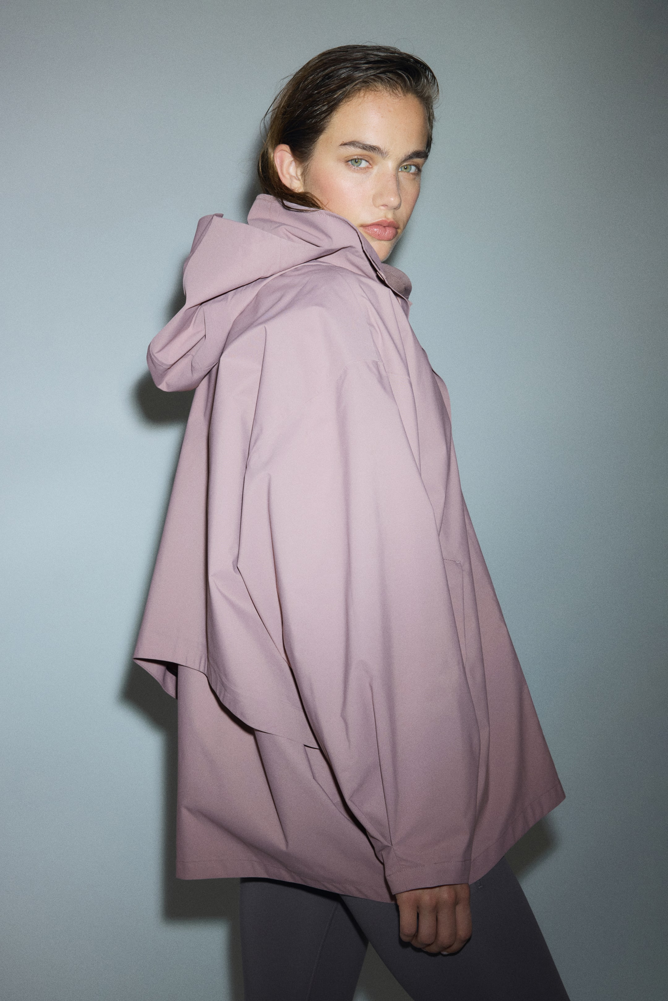 Visualizza immagine più grande: Un poncho color malva con cappuccio viene presentato da un'angolazione laterale-posteriore. Presenta una silhouette ampia e fluente con maniche larghe e drappeggiate e un tessuto liscio e non testurizzato.