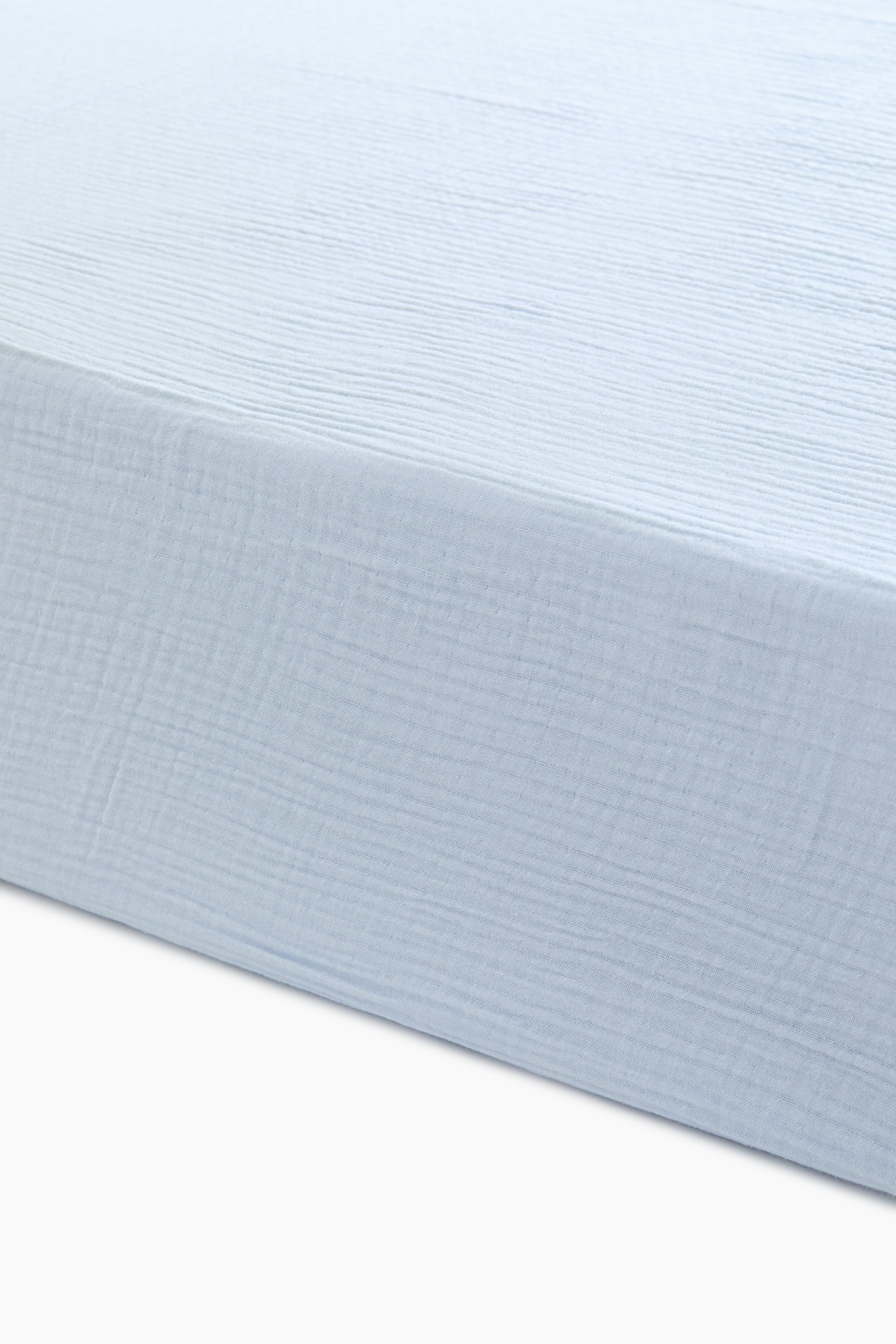 View larger image: A light blue cot fitted sheet, made of soft cotton muslin, appears stretched and fitted. Its top surface and front side exhibit the characteristic crinkled texture of muslin fabric.