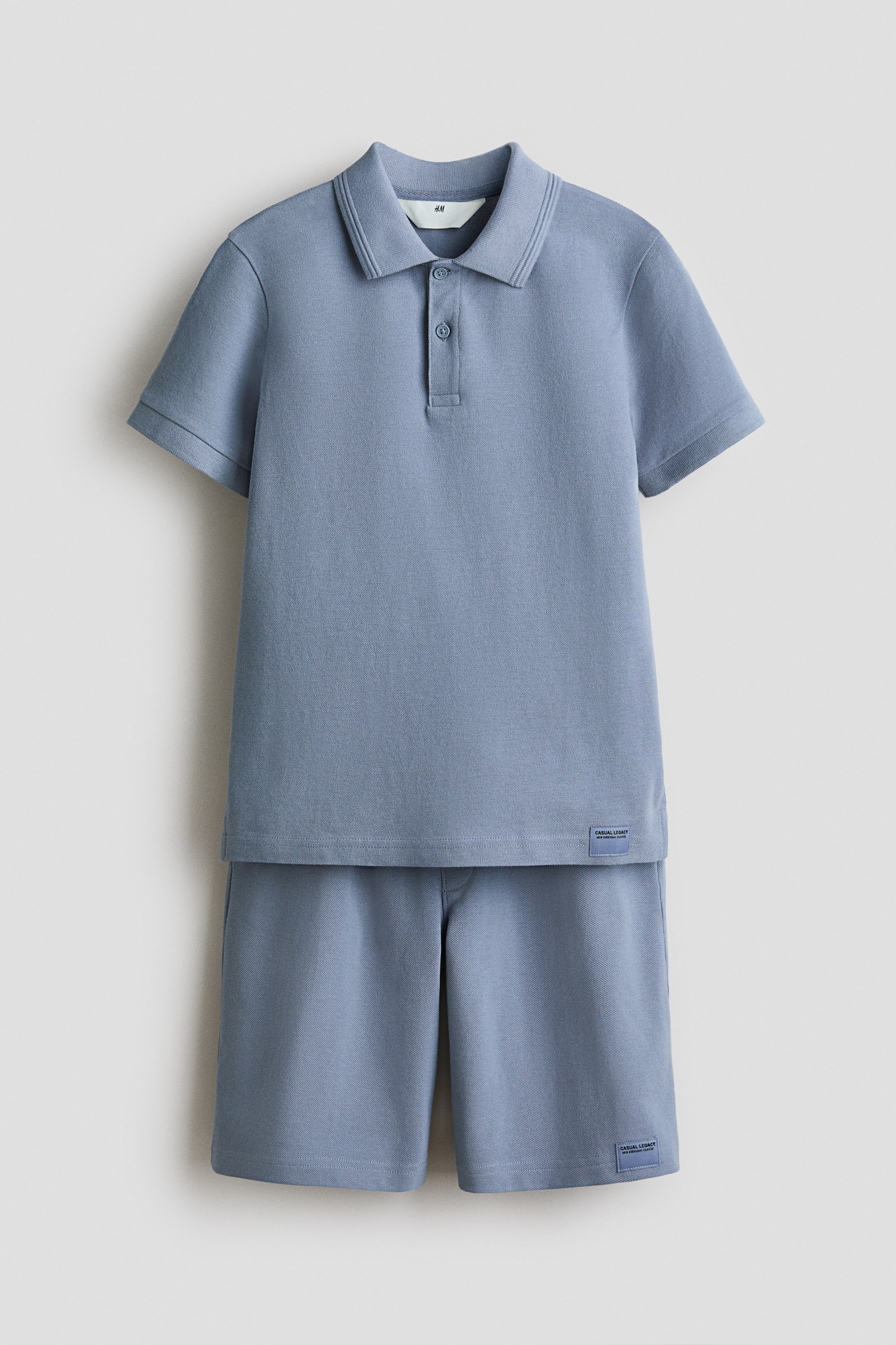 Ingrandisci l'immagine: Un completo in maglia jersey azzurro chiaro a due pezzi è presentato frontalmente, composto da una polo a maniche corte e pantaloncini coordinati. La polo presenta un colletto a coste e un'abbottonatura a due bottoni, e entrambi i pezzi mostrano una piccola etichetta rettangolare tessuta con la scritta 'Casual Legacy' vicino all'orlo.