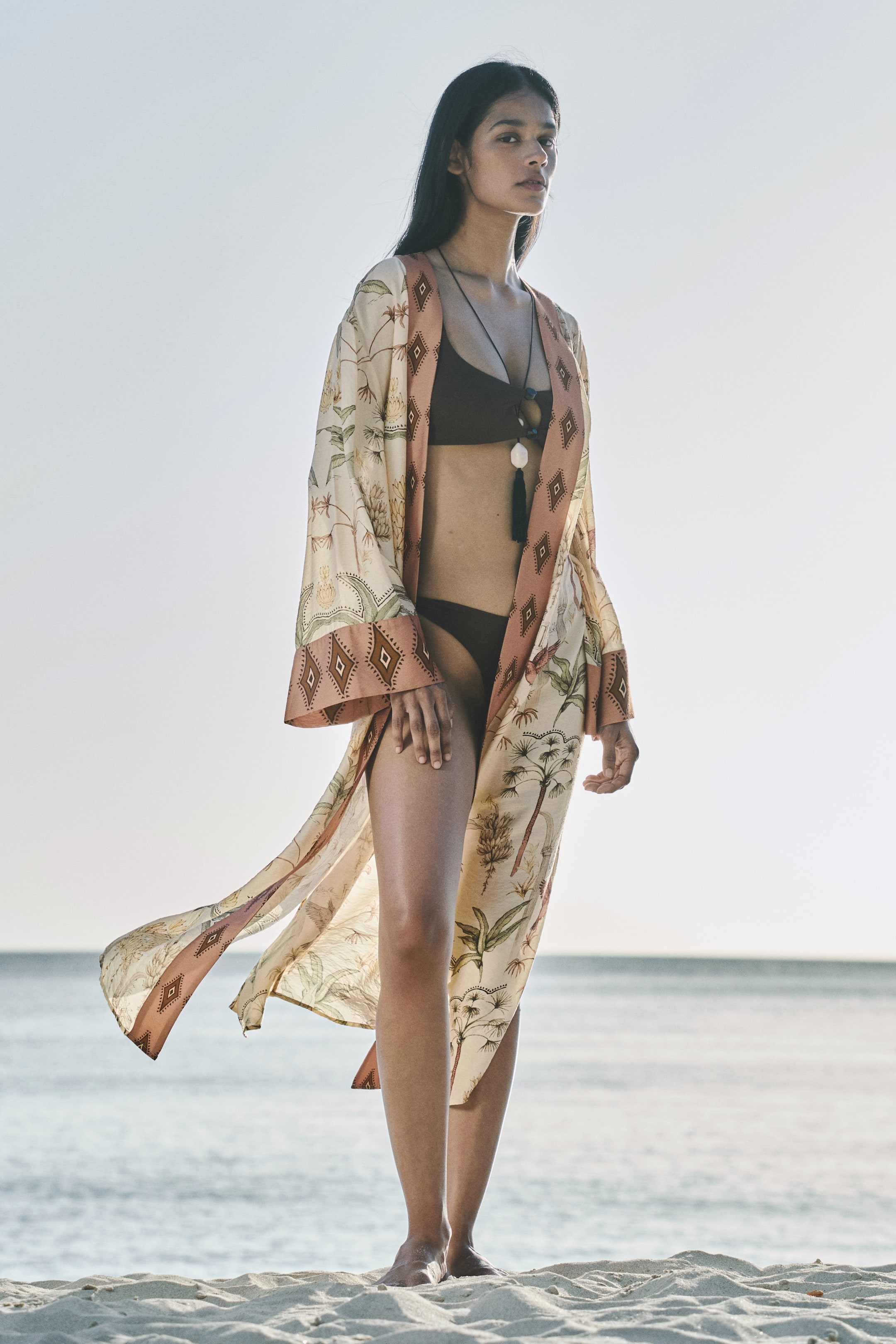 View larger image: A long, flowing kimono-style cover-up is worn open, facing forward. It showcases a light beige fabric adorned with botanical motifs, bordered by a reddish-pink band featuring a diamond geometric pattern. The cover-up drapes over a dark brown bikini top and bottom.