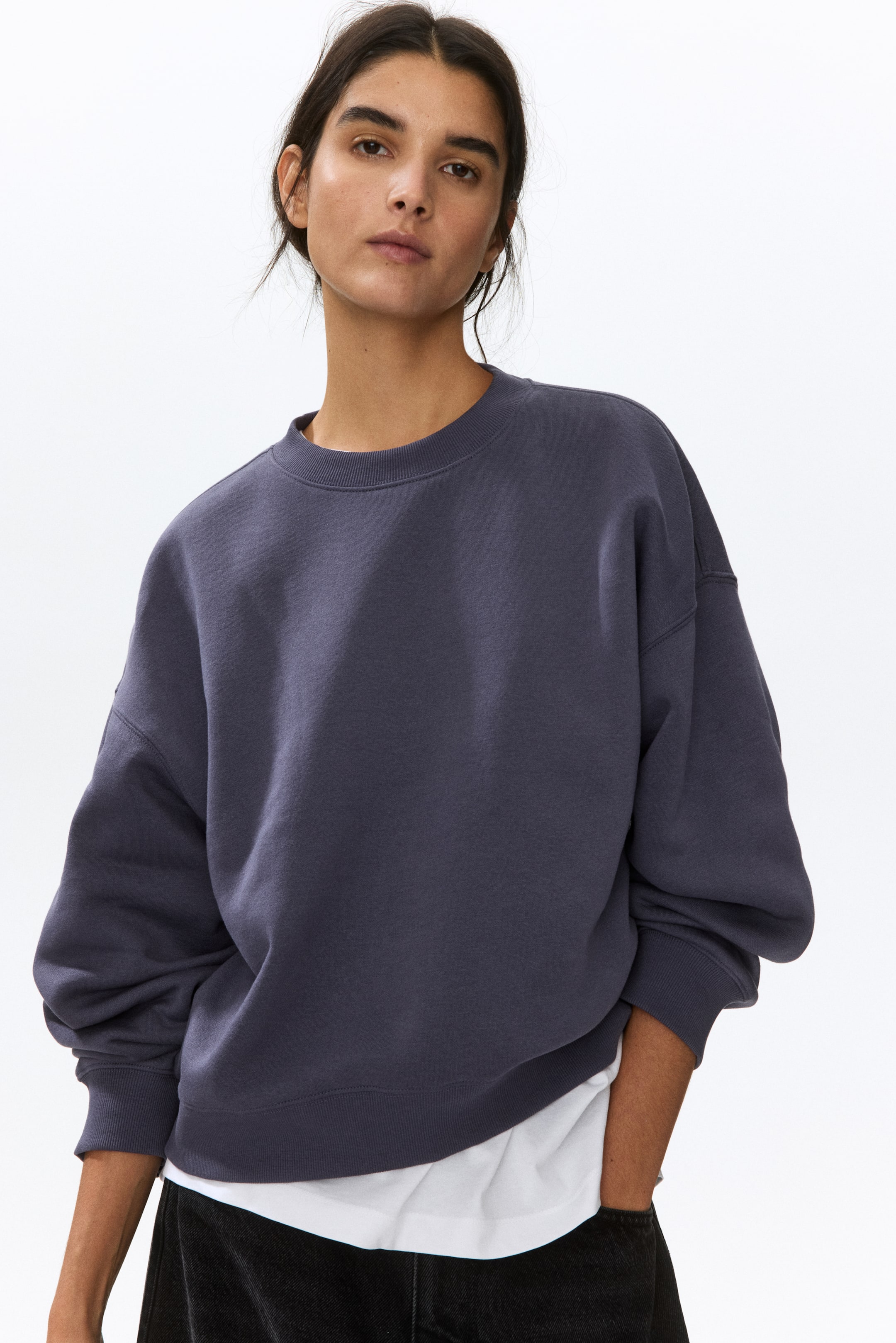 View larger image: A dark gray oversized sweatshirt is displayed from a front-facing angle, featuring a ribbed crew neck, dropped shoulders, and ribbed cuffs and hem, with a white t-shirt layered beneath its hem.