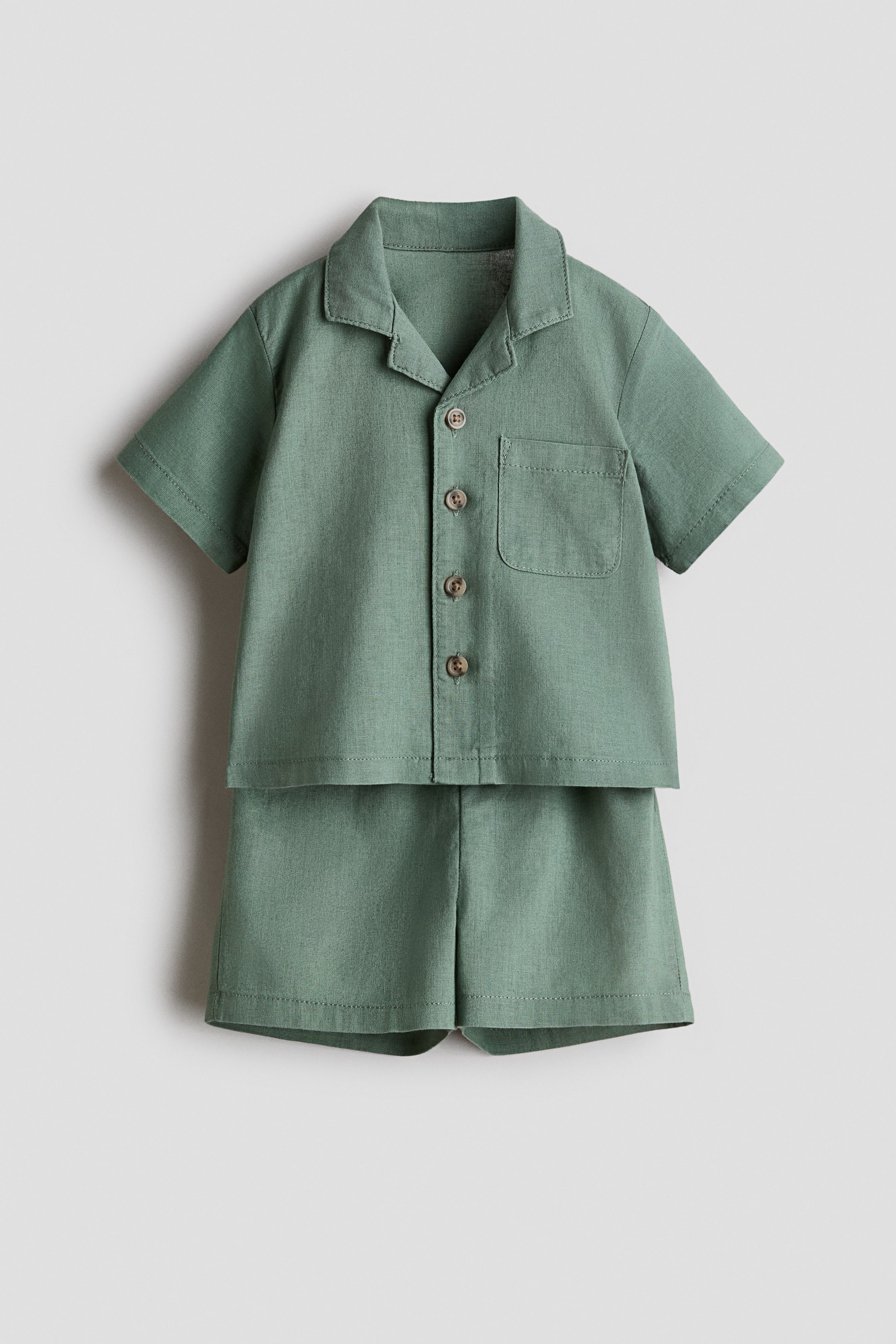 View larger image: A front-facing green set features a short-sleeved shirt and matching shorts, both showcasing a linen and cotton weave. The shirt highlights a resort collar, wood-look buttons down the front, a chest pocket, and dropped shoulders, while the shorts present sewn-in turn-ups at the hems.