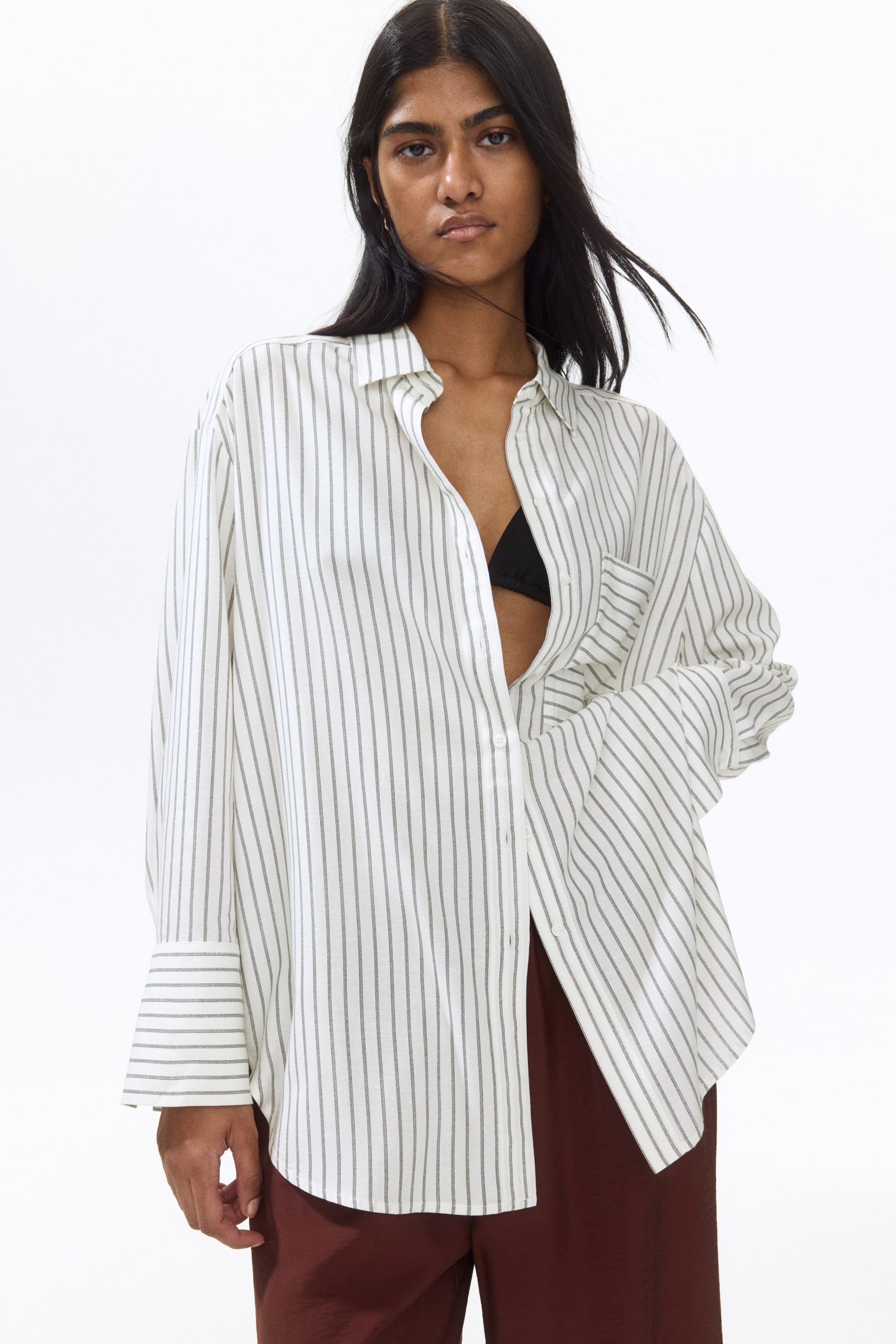 View larger image: The model is wearing an oversized white shirt adorned with thin grey vertical stripes. It features a classic collar, an open chest pocket, dropped shoulders, and long sleeves with cuffs. The shirt is partially unbuttoned, subtly revealing a black top underneath. Its straight-cut hem with rounded side vents elegantly drapes over brown trousers.