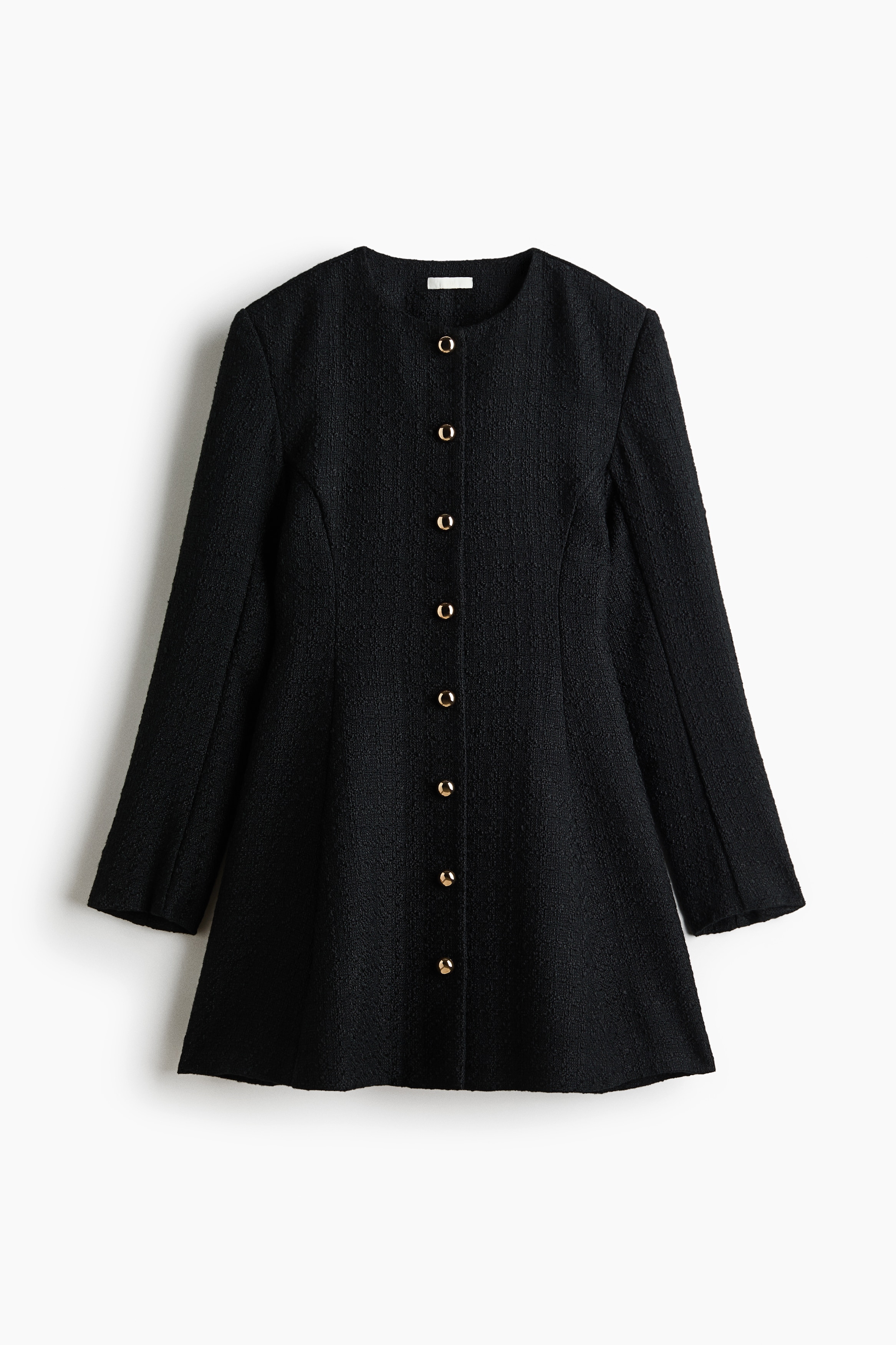 A black textured long-sleeve dress featuring a round neckline, structured shoulders, and a front closure with metallic dome buttons is being worn by the model. The dress is styled with dark rectangular sunglasses as the model stands with arms crossed.