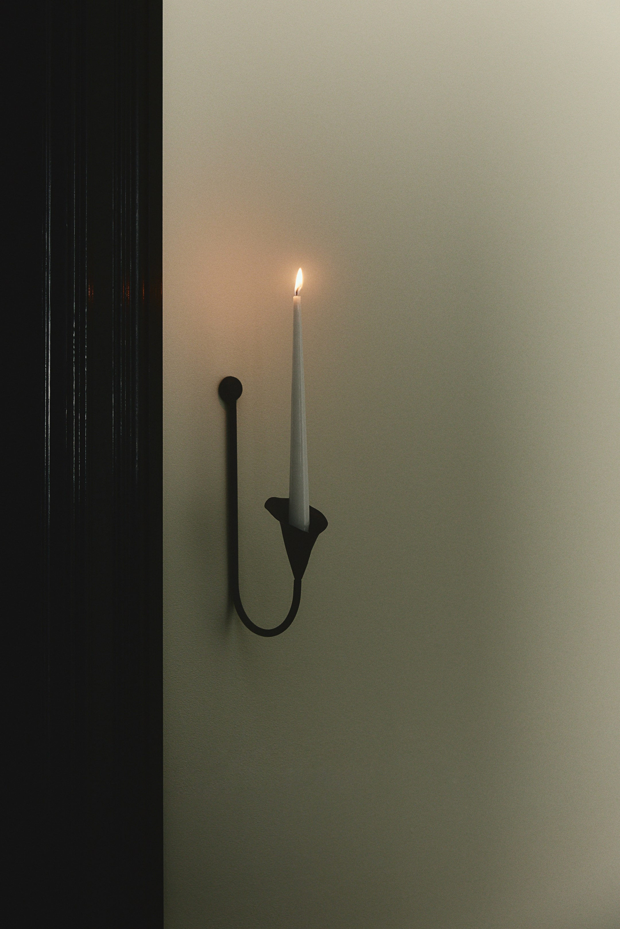 View larger image: A black metal wall sconce is shown in profile, featuring a U-shaped rod that curves from a circular mount into a flower-shaped candle holder. A tall, lit white taper candle is resting upright inside the flower-shaped cup.
