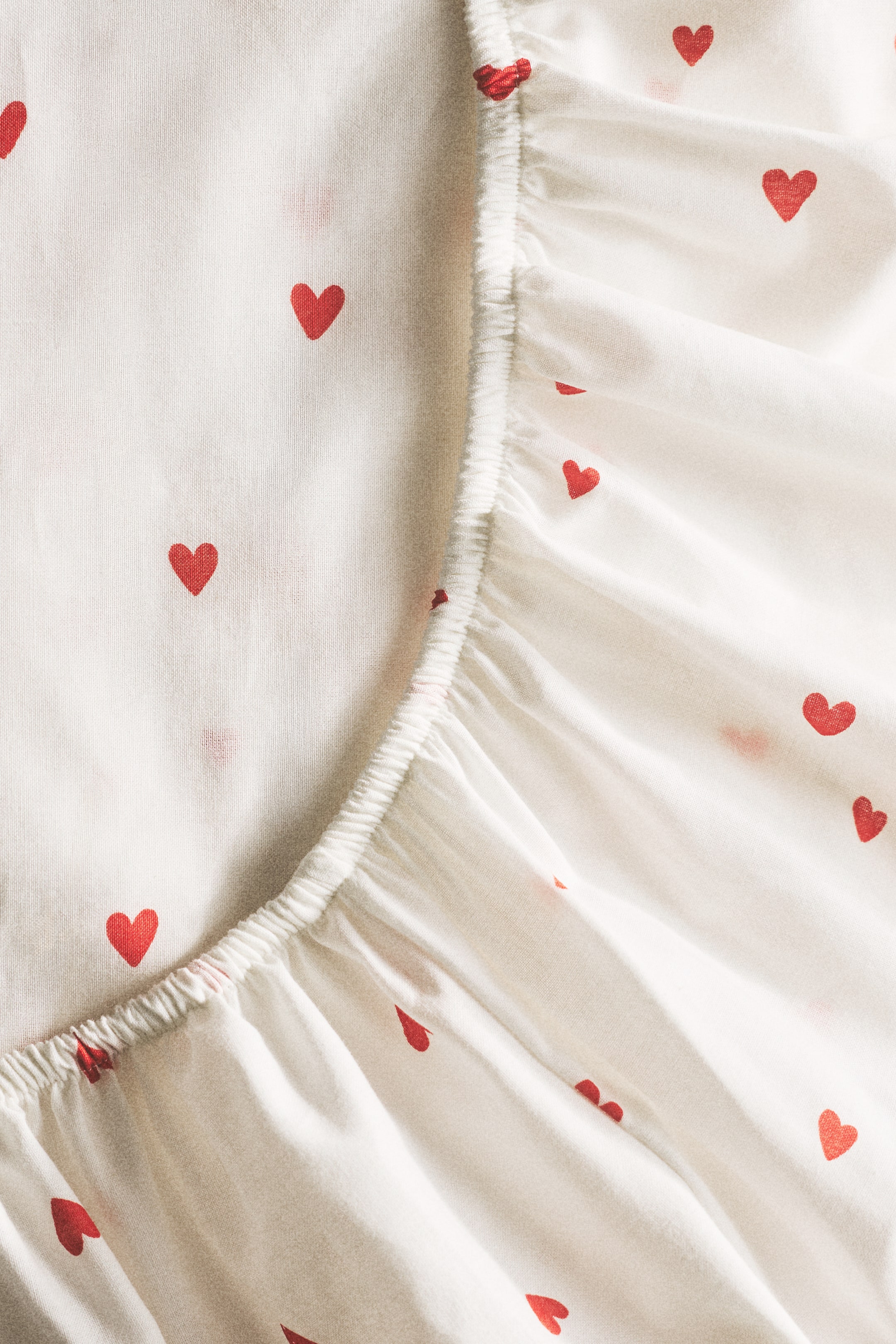 View larger image: The fitted sheet fabric features a pattern of small red hearts on an off-white background. The fabric showcases its elasticized, gathered edge, creating soft folds.
