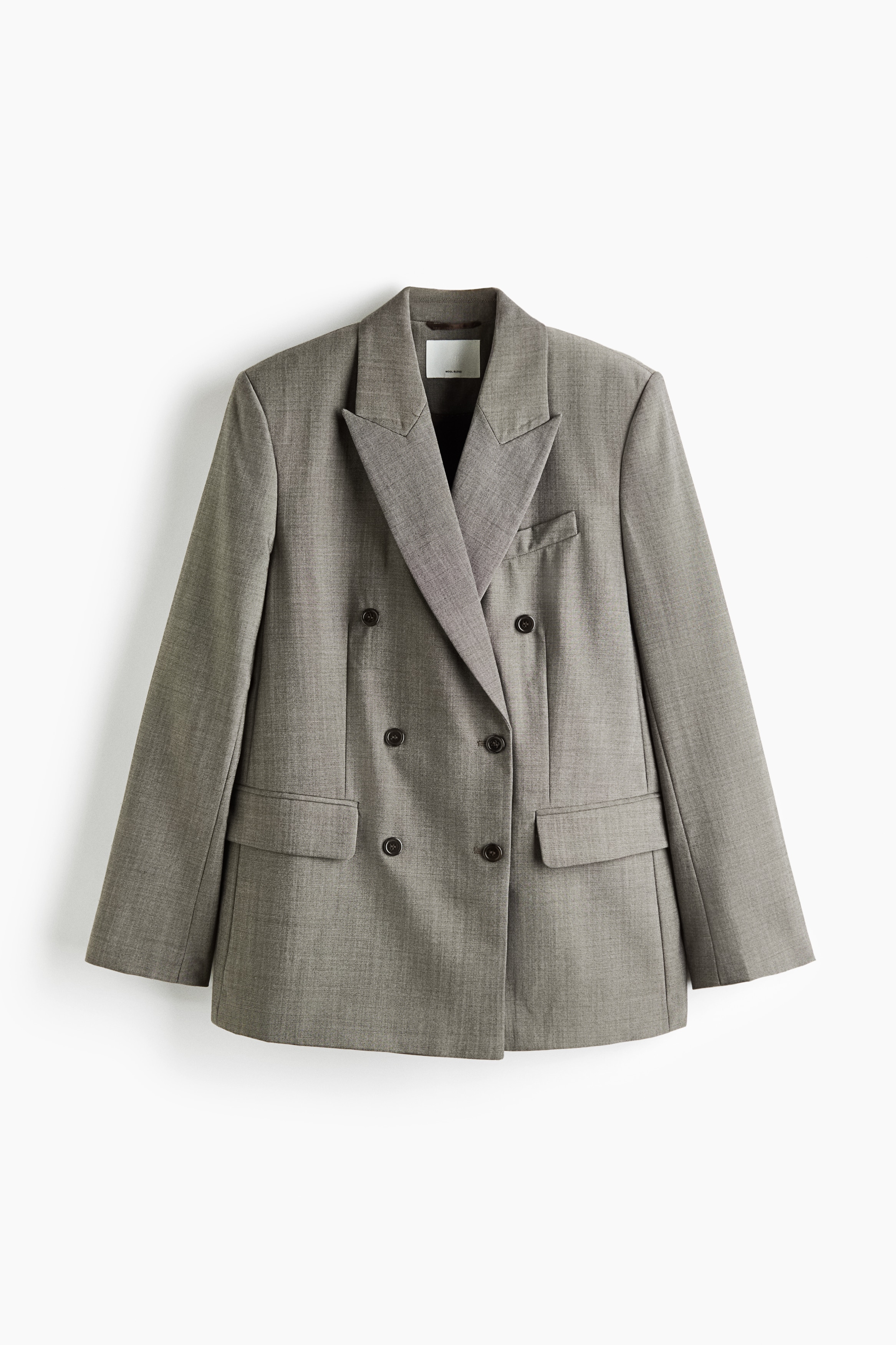 La modelo lleva puesto un blazer gris de corte oversize, cruzado y confeccionado en un tejido de mezcla de lana. El blazer presenta amplias solapas de pico, un bolsillo de pecho ribeteado y botones oscuros en el delantero.