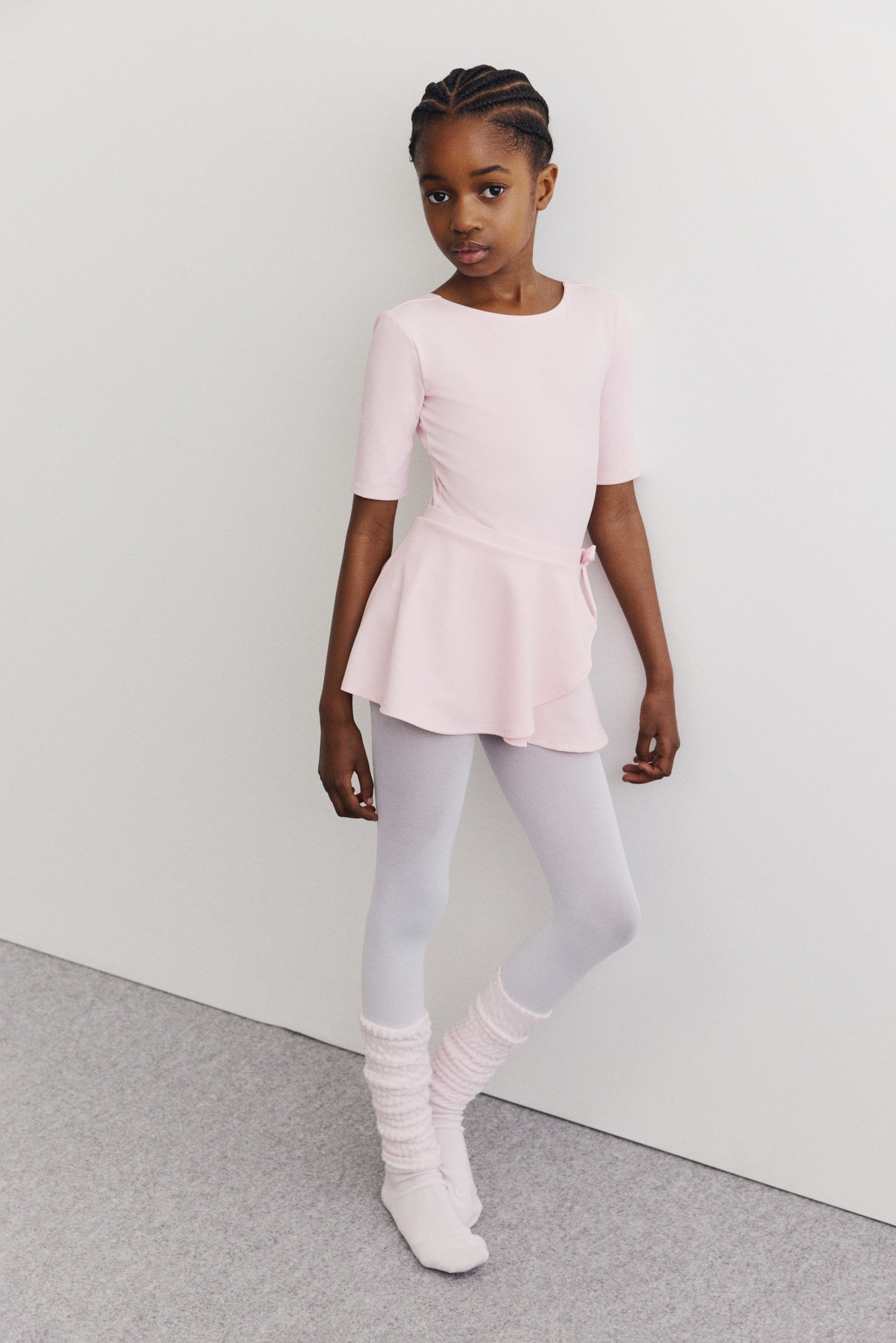 Vezi imaginea mai mare: Este prezentat un costum de balet pentru copii, compus dintr-un leotard roz pal cu mâneci scurte și decolteu rotund, o fustă petrecută roz pal cu legătură laterală, dresuri albe lungi și încălzitoare pentru gambe roz pal, plisate, purtate peste șosete de balet roz pal.