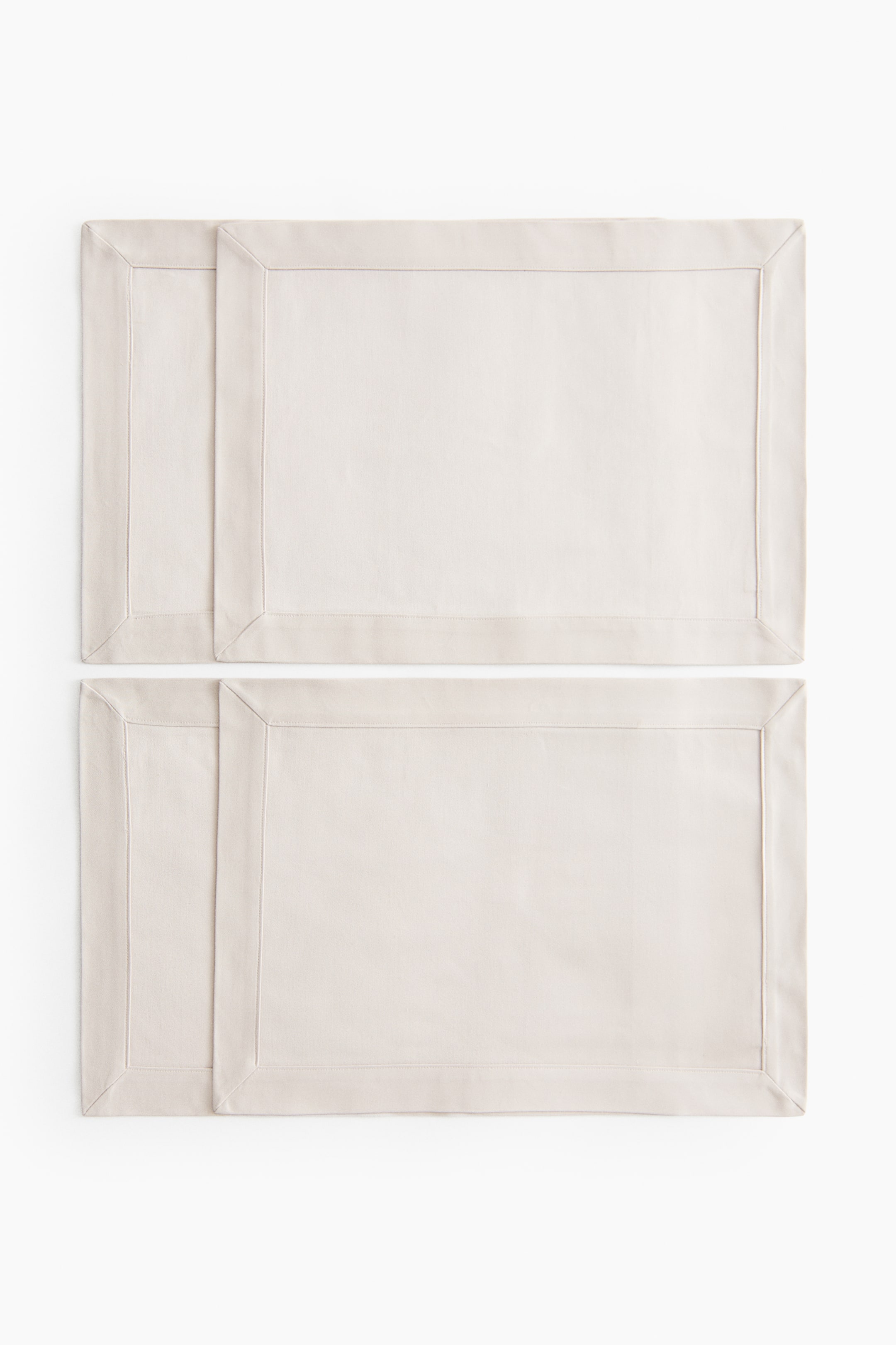 View larger image: Four light cream rectangular placemats feature a wide stitched border. They are arranged in two pairs, with the left placemat in each pair slightly overlapping the one on the right.