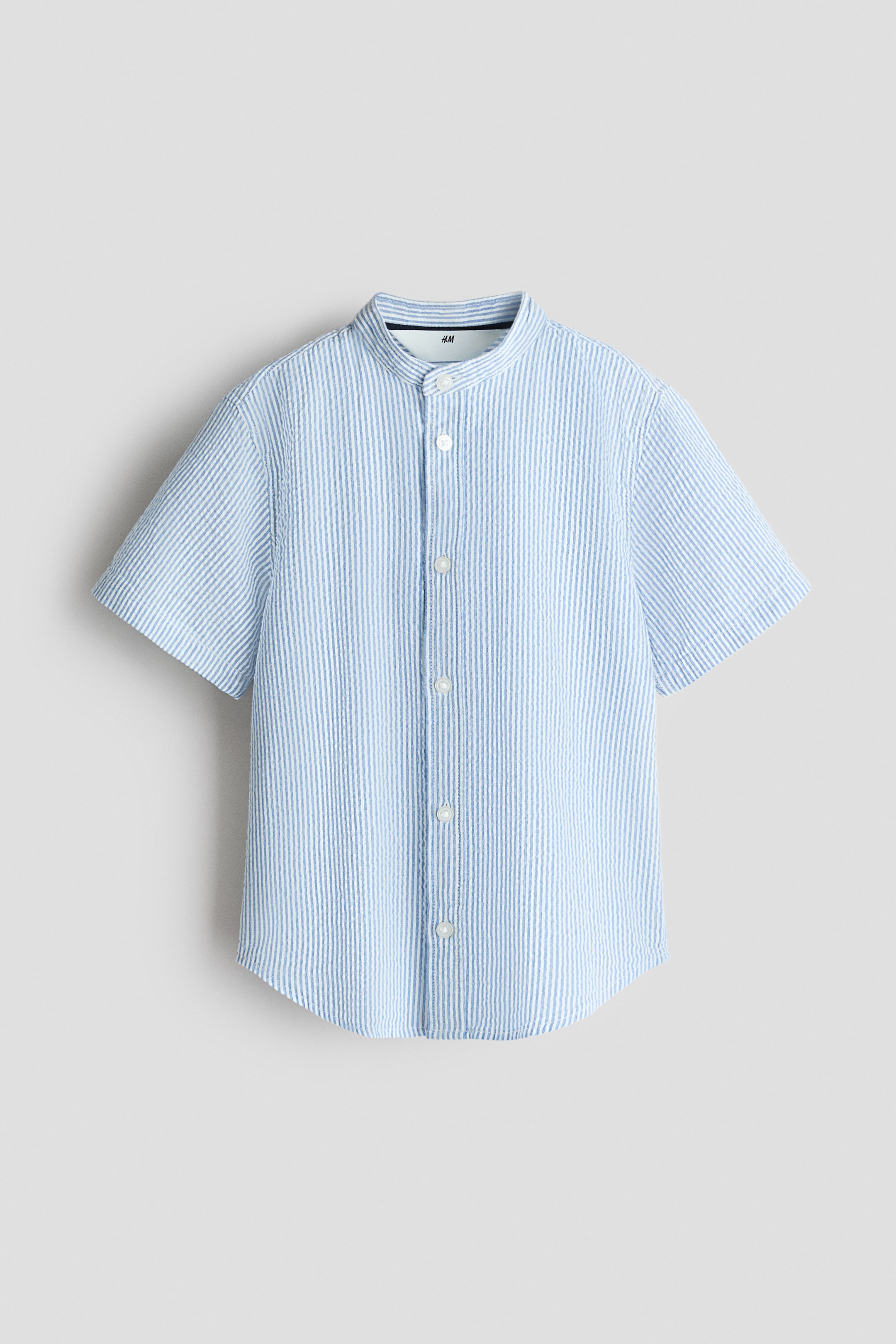 View larger image: "A short-sleeved shirt is facing front, showcasing light blue and white vertical seersucker stripes. It comes with a band collar, a full-button front closure with white buttons, and a rounded hem."