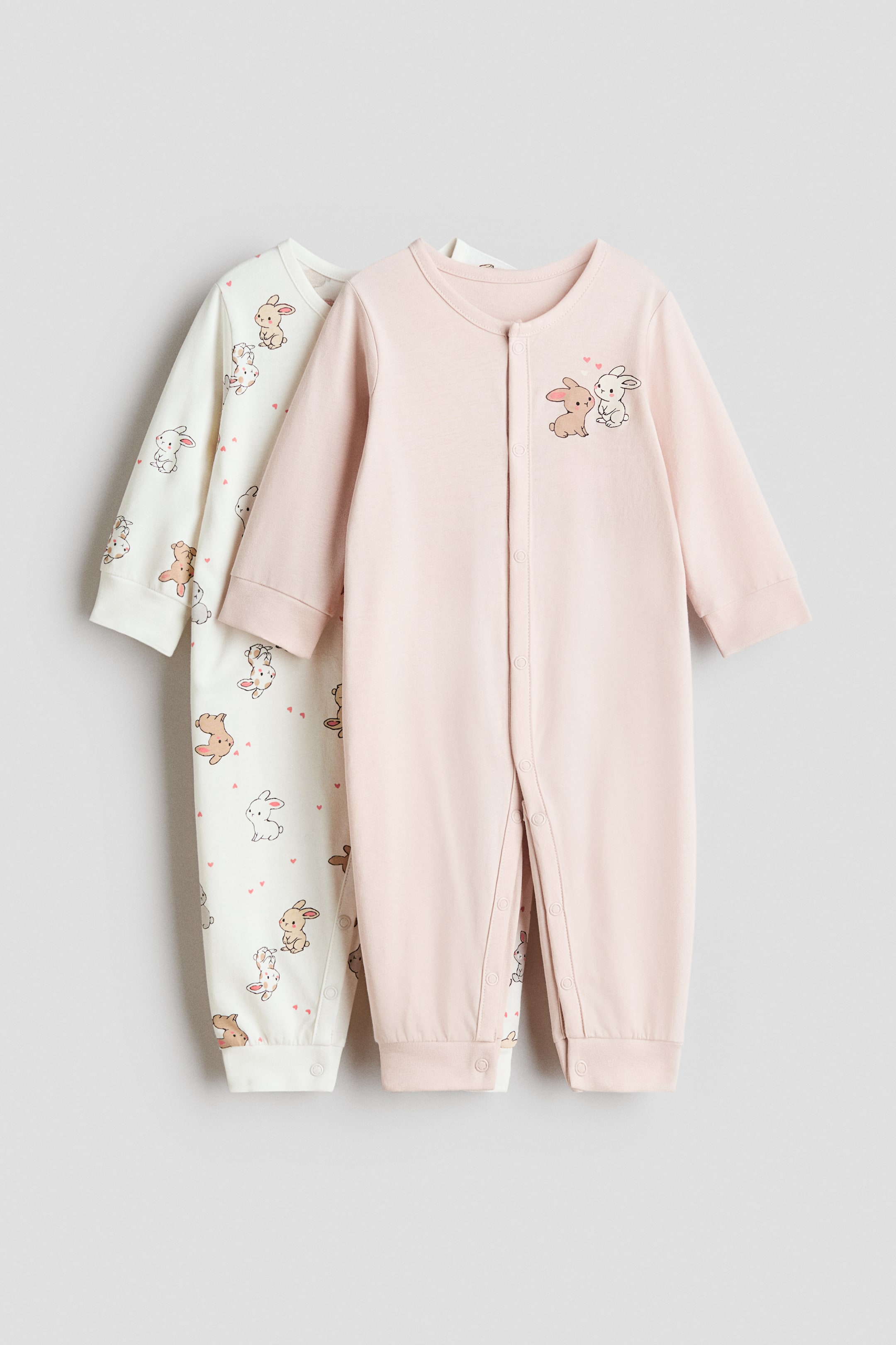 拡大画像を表示: 2つのスリープスーツは前面を見せています。ピンクのスリープスーツは胸にうさぎのイラストが描かれ、オフホワイトのスリープスーツは全面にうさぎとハートのプリントがあります。両方のスリープスーツは、長袖、リブ付きのカフスと裾、そして前から脚にかけて伸びるスナップボタンを特徴としています。