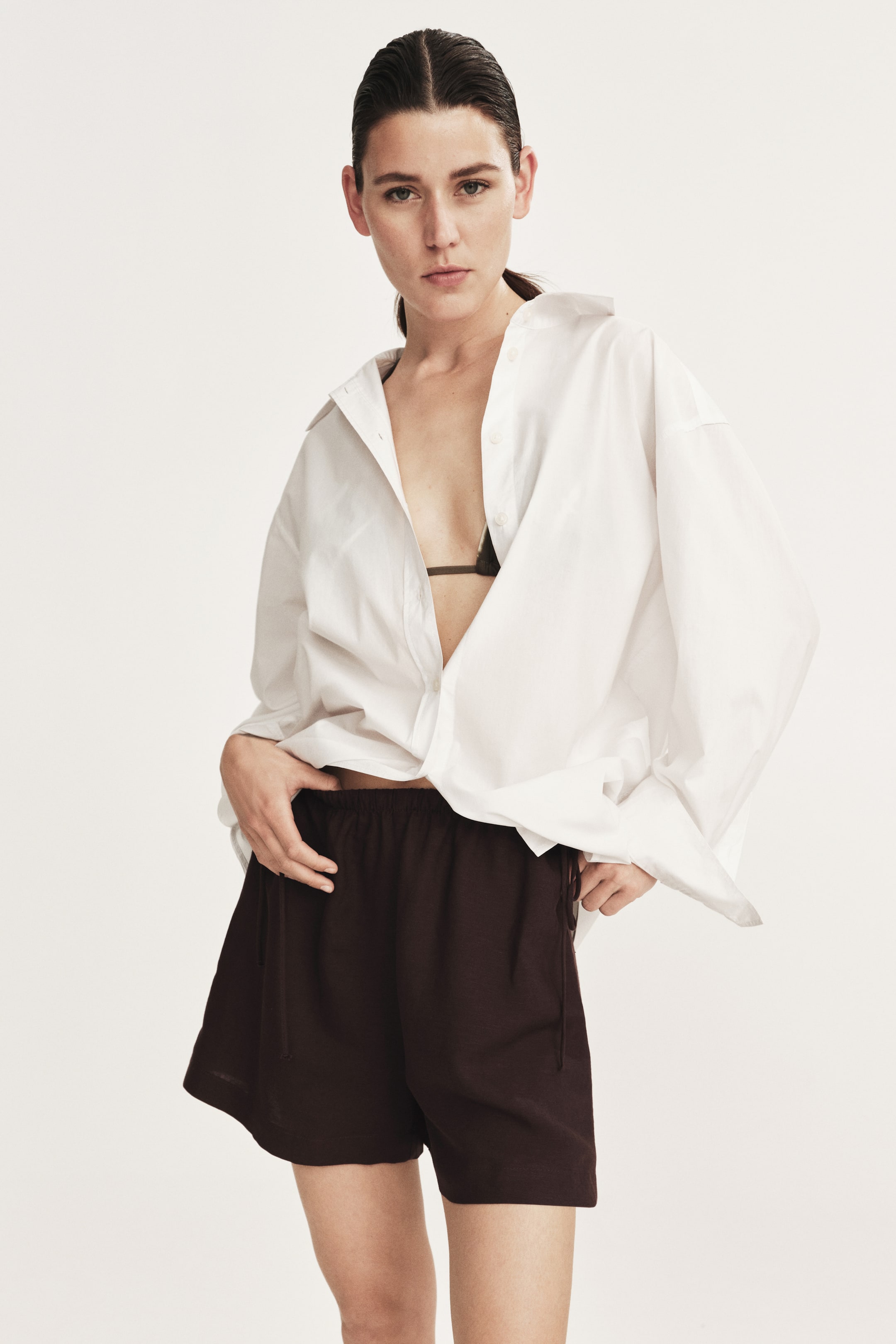 View larger image: A white oversized button-down shirt with a pointed collar is being worn open over a dark triangle-style bra, with its long sleeves and front partially tucked into dark brown loose-fitting shorts. The shorts feature an elasticated waistband with a visible drawstring, extending to mid-thigh.