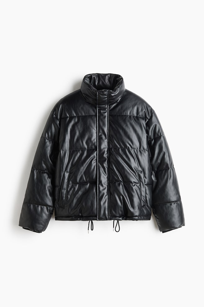 A boxy black puffer jacket in a coated, leather-like fabric is presented in a front-facing view, featuring a high stand collar, horizontal quilting, and dropped shoulders. It also features a concealed front closure, welt side pockets, and adjustable drawstring cords hanging from the hem. - 1