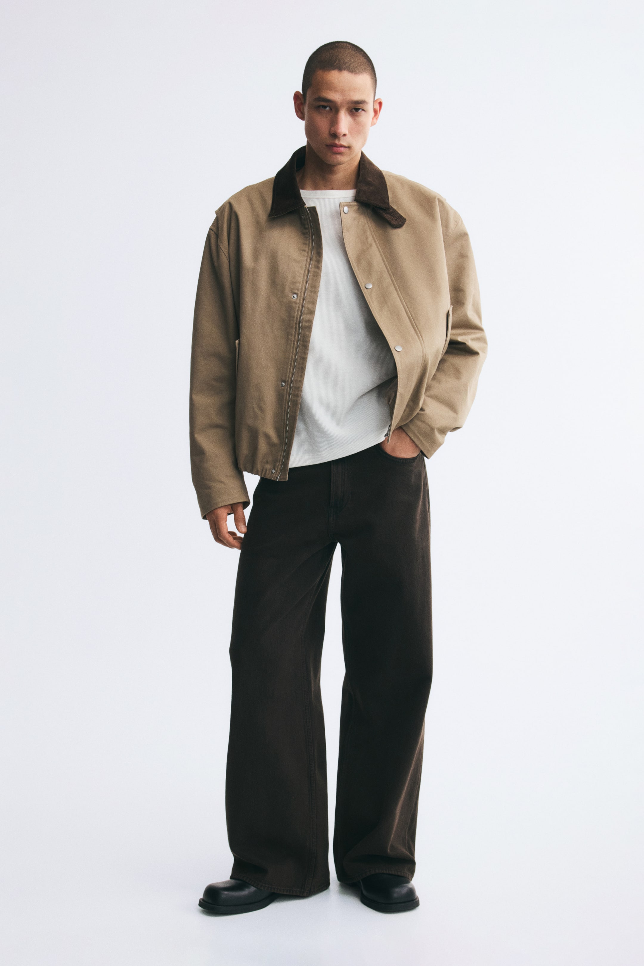 View larger image: Dark brown, wide-leg denim jeans with a loose fit are worn by the model, who is standing with one hand tucked into a front pocket. The jeans are styled with a white T-shirt, a tan jacket with a brown collar, and black shoes.