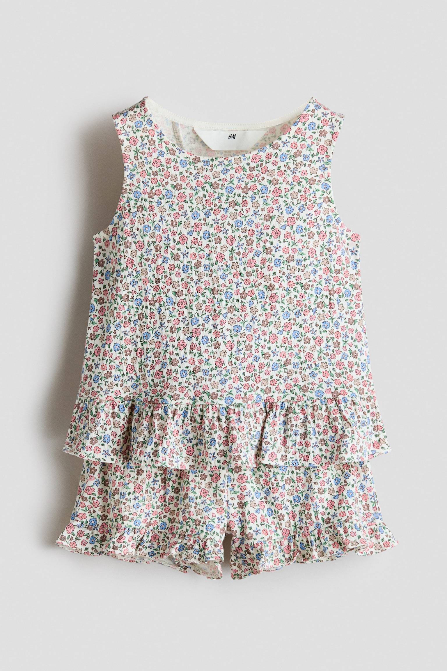 A front-facing view of a children’s sleeveless top and shorts set featuring an all-over ditsy floral pattern in pink, blue and green on a cream-coloured fabric. The sleeveless top has a round neckline and a tiered ruffled hem, layered over matching shorts with ruffled leg openings.
