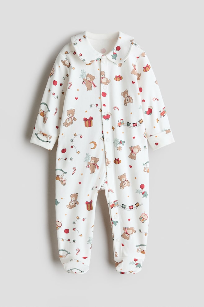 A collared cotton sleepsuit is shown from a front-facing view, featuring a rounded Peter Pan collar, long sleeves with folded cuffs, and integrated feet. The sleepsuit features an all-over festive print of teddy bears, toy trains, and wrapped gifts, with a snap-button closure running down the centre front. - 1