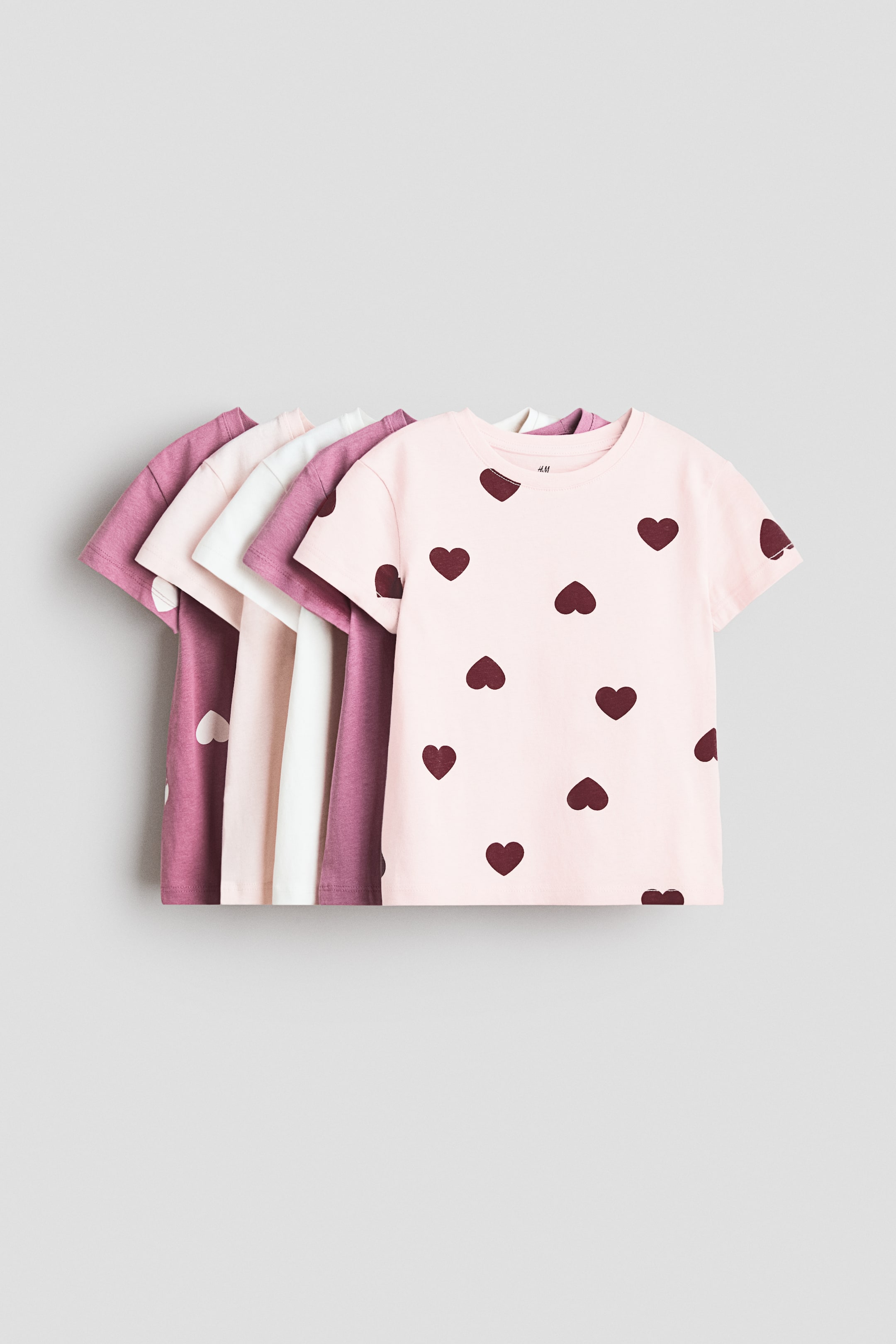Ver imagem maior: As T-shirts em jersey de algodão macio apresentam uma gola redonda com acabamento em canelado e ombros ligeiramente caídos, com as suas frentes voltadas para a frente. A T-shirt mais à frente exibe um padrão de corações escuros repetidos sobre um fundo rosa claro, enquanto T-shirts de cores sólidas em rosa, branco e roxo são parcialmente visíveis em camadas por trás dela.