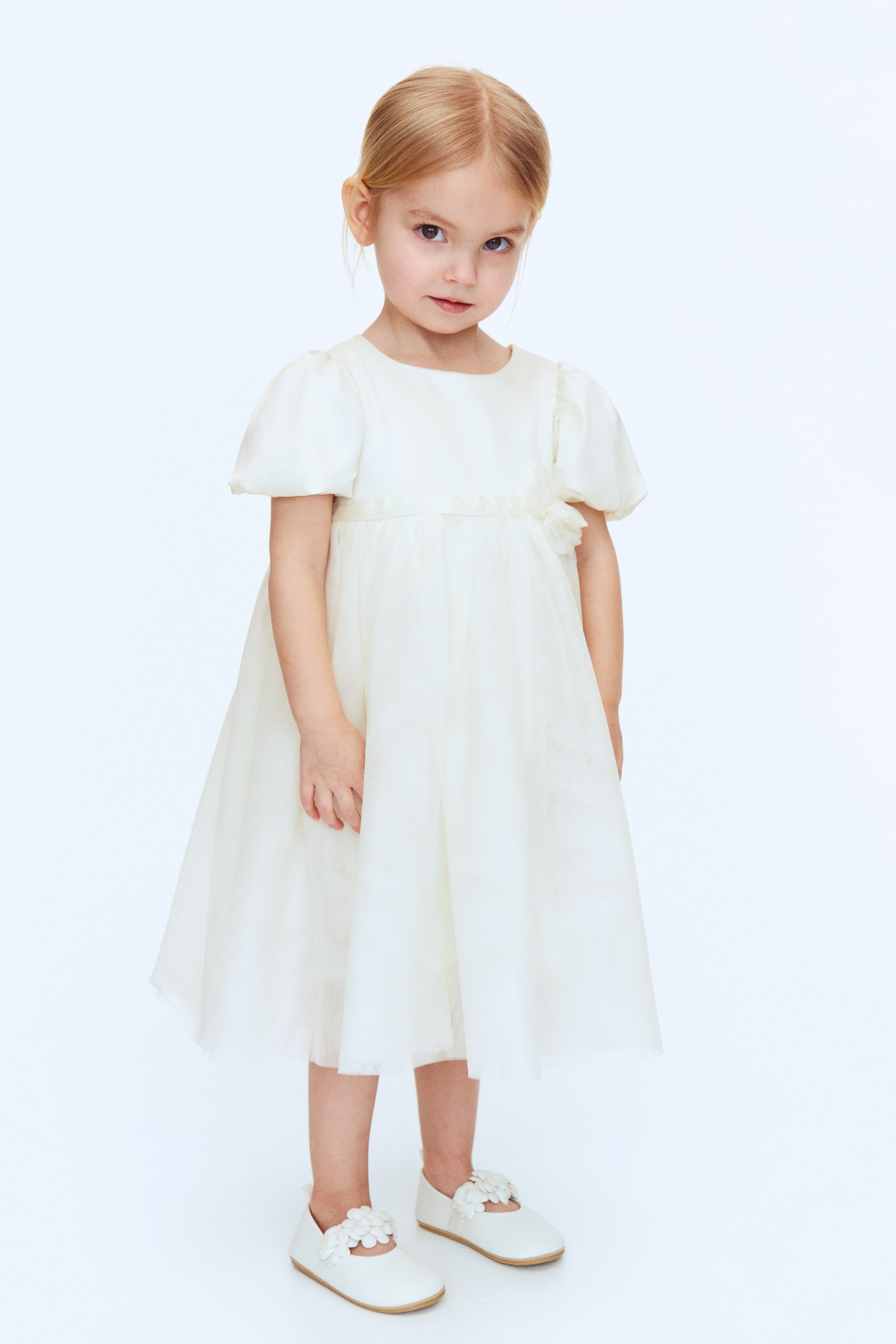 View larger image: An off-white short-sleeve dress features a round neckline and puff sleeves. A satin band accentuates the empire waist, adorned with a floral rosette on the wearer's left, as the flowing skirt falls below the knee.