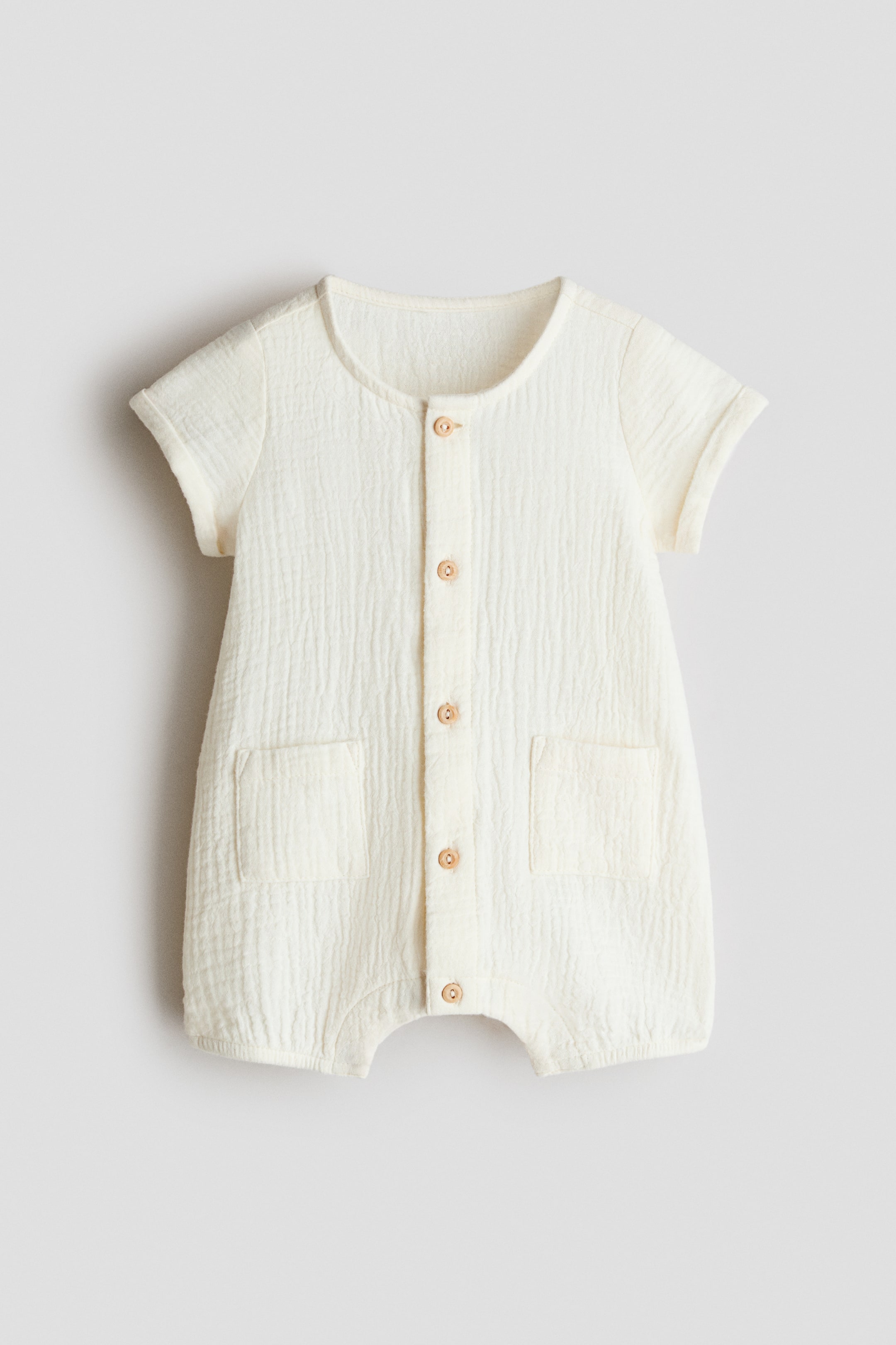Agrandir l'image: Une combinaison crème en mousseline de coton froissée est présentée de face, avec un col rond et une patte de boutonnage avant avec six boutons clairs. Elle présente également des manches courtes avec revers cousus, deux poches plaquées à l'avant et des jambes courtes avec des ourlets élastiqués.