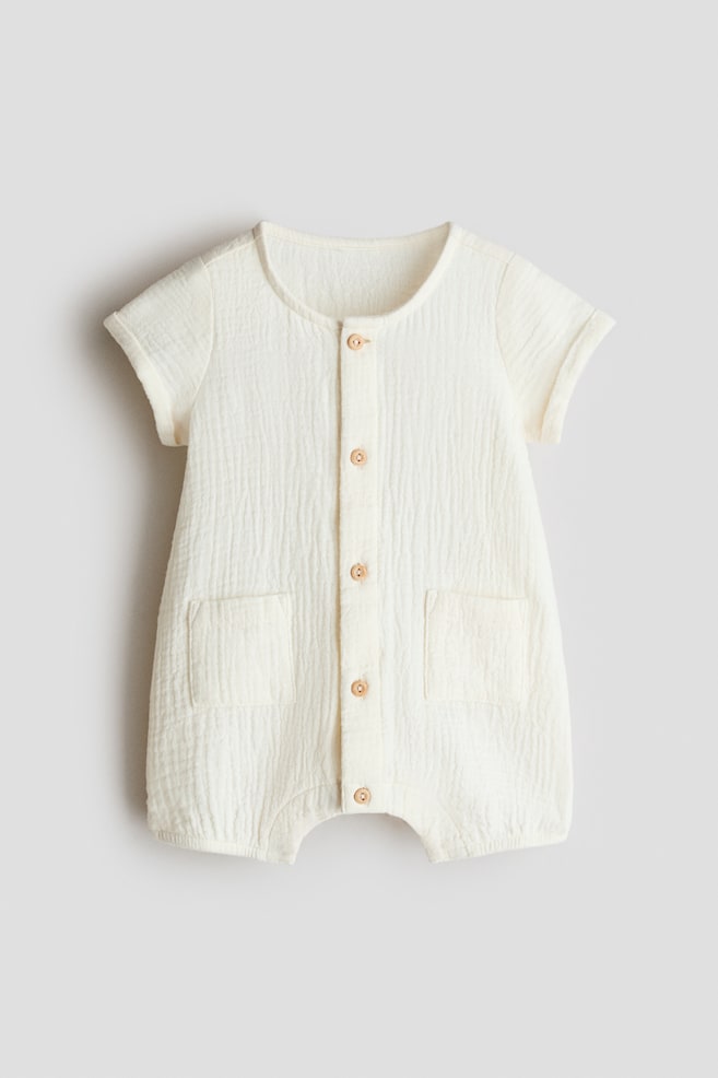 A cream-colored romper suit in crinkled cotton muslin is facing front, featuring a round neckline and a front button placket with six light buttons. It is also showing short sleeves with sewn-in turn-ups, two patch pockets on the front, and short legs with elasticated hems. - 1