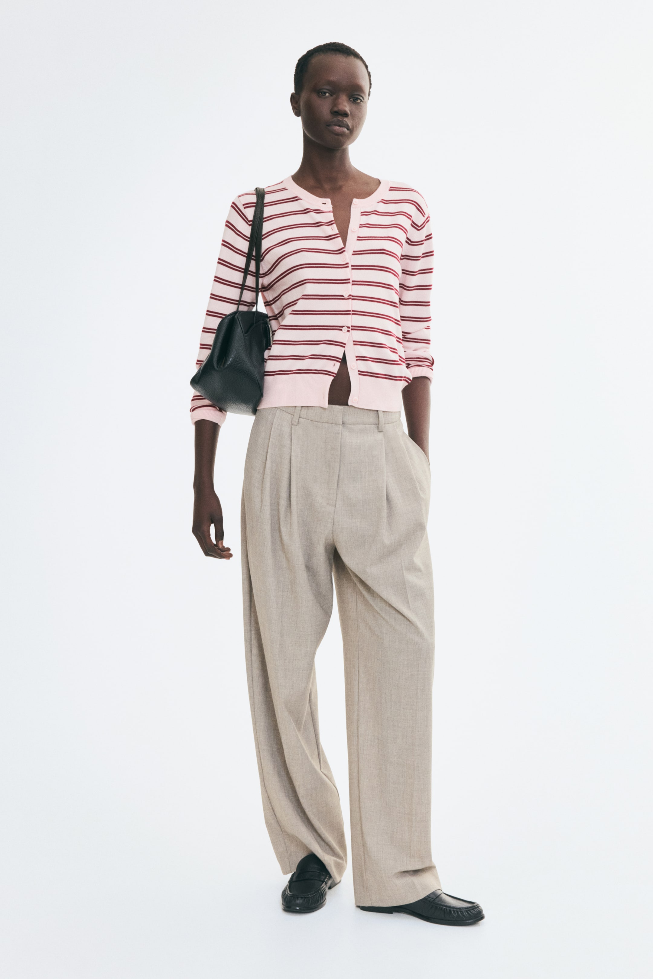 View larger image: The model is wearing a light pink and red striped knit cardigan, featuring a round, rib-trimmed neckline, long sleeves, and a button-front closure left partially open at the hem. The cardigan is styled with beige wide-leg trousers and a black shoulder bag.