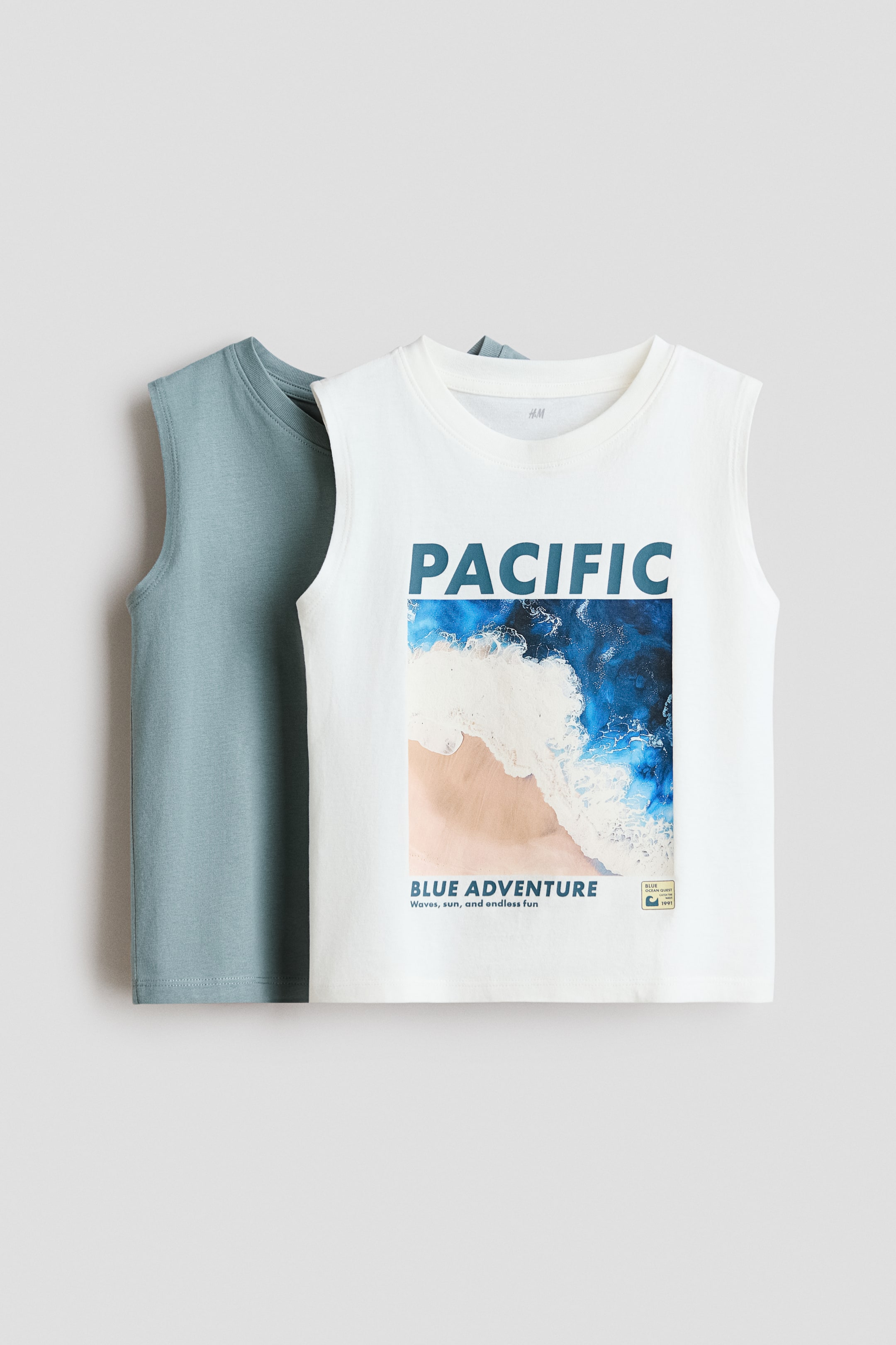 View larger image: The front tank top is an off-white, round-neck, sleeveless top showcasing a prominent blue 'PACIFIC' graphic above a rectangular aerial beach scene with blue and white waves meeting a sandy shore, accompanied by the smaller text 'BLUE ADVENTURE Waves, sun, and endless fun'. A second teal tank top is partially visible behind it.